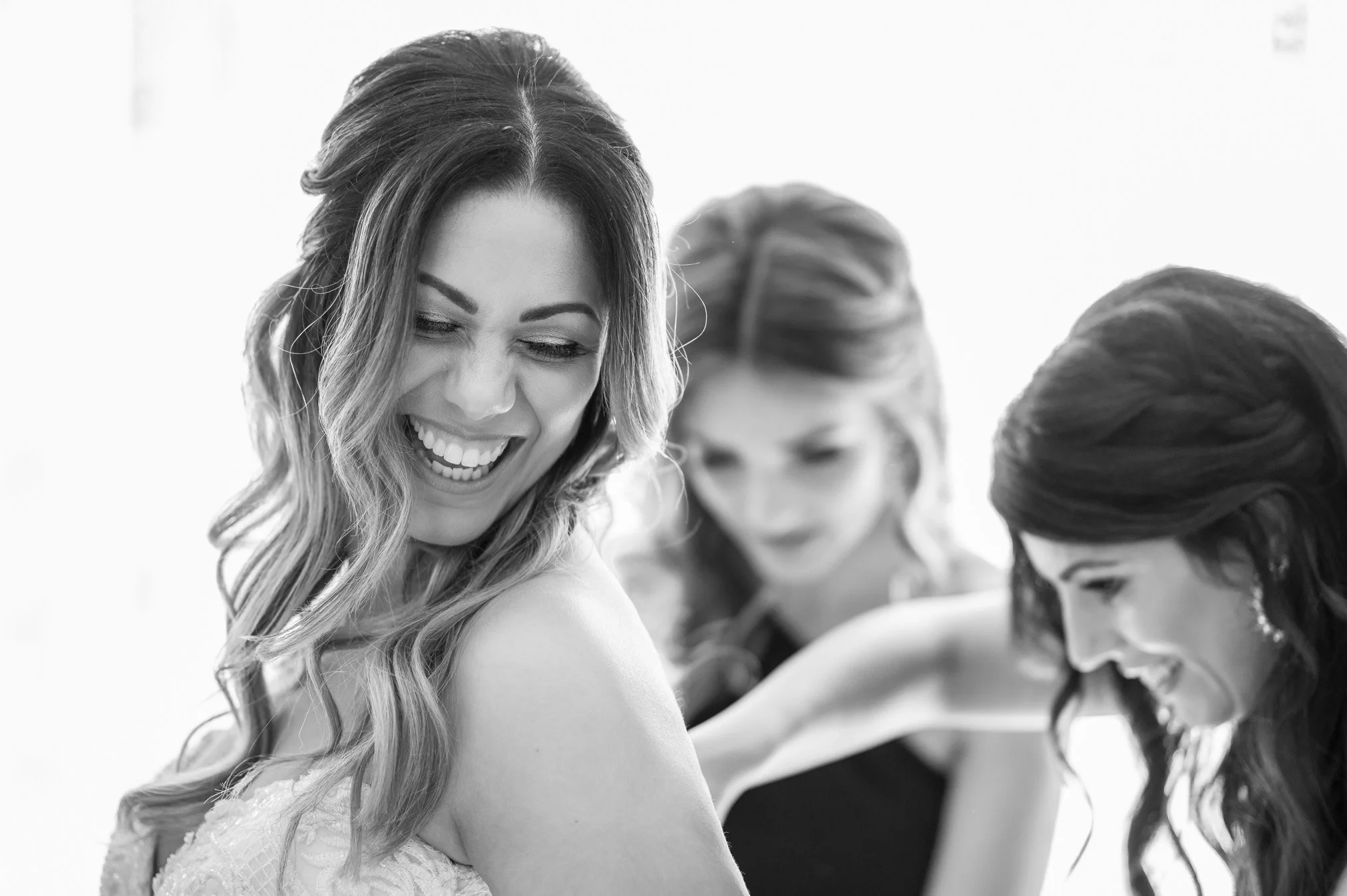 Three women, with wavy hair, smiling and looking down, in a candid black-and-white photo.