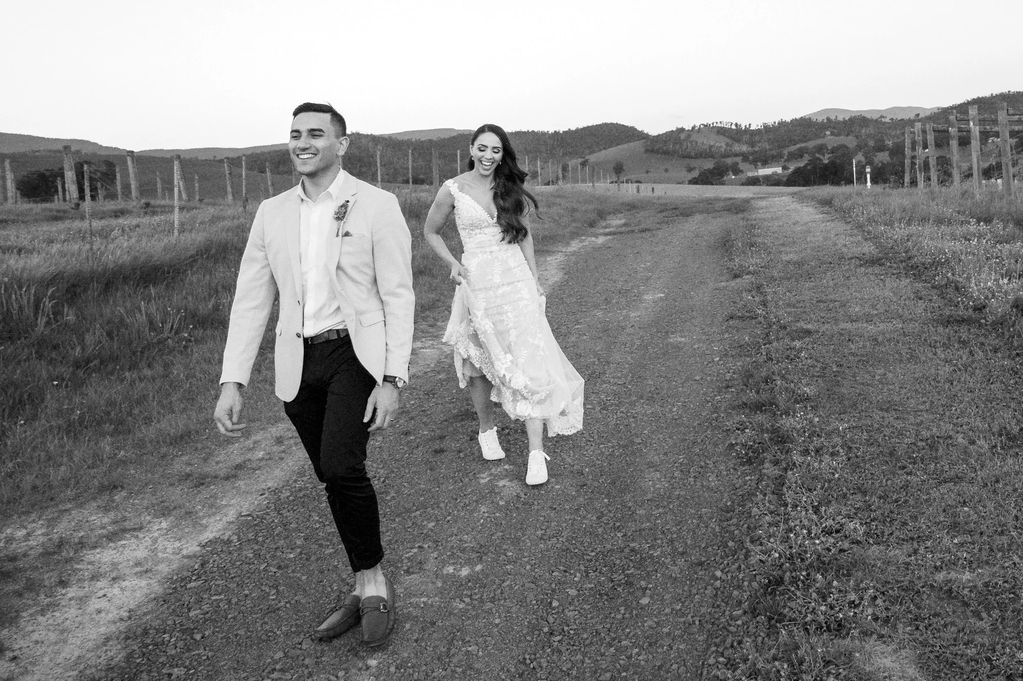 A black and white photo of a happy couple, dressed in wedding attire, walking on a dirt path in a rural landscape with hills in the background. The groom wears a light-colored suit jacket, white shirt, and dark pants, and the bride wears a lace weddi