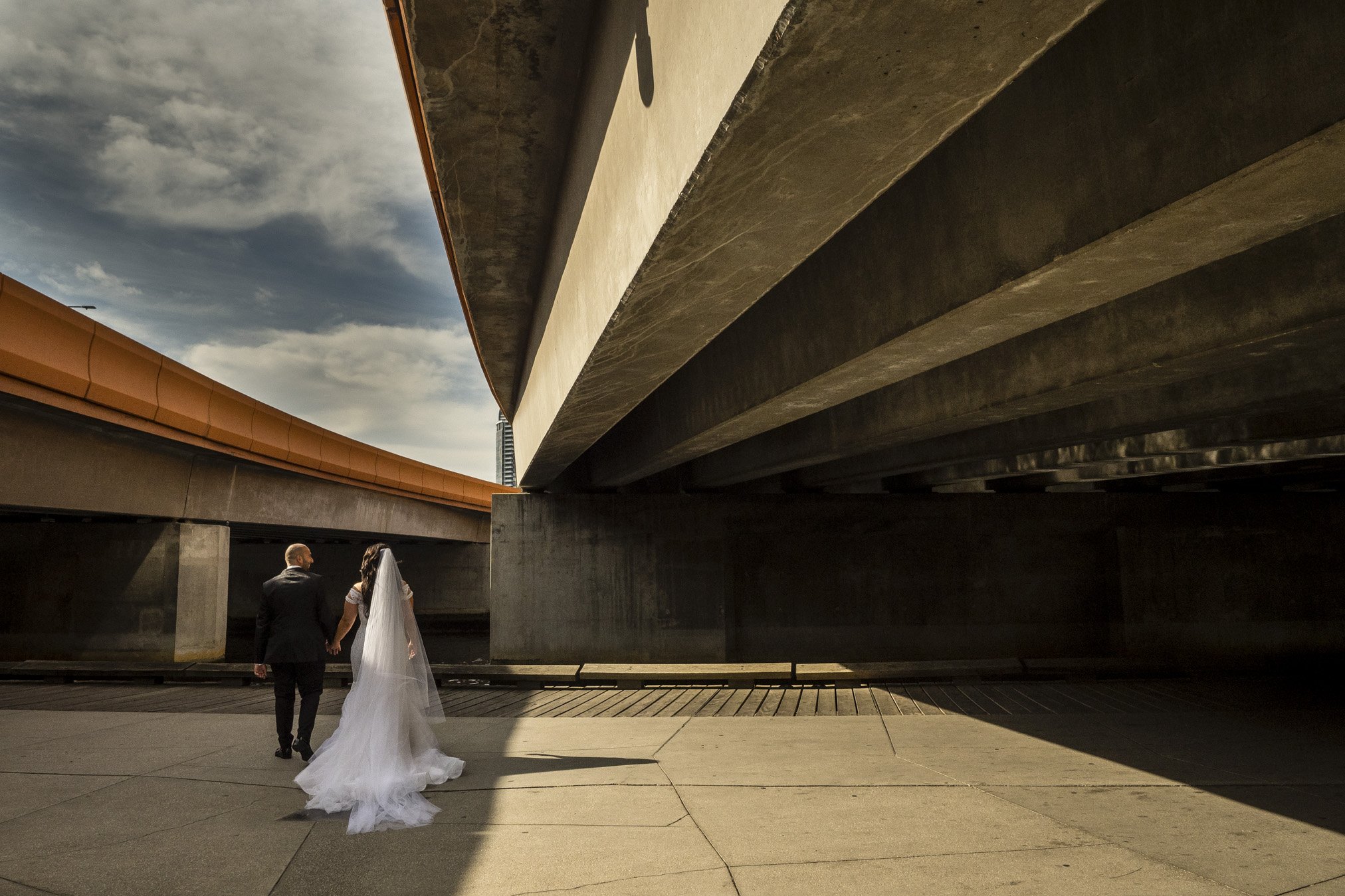 A bride and groom walking hand in hand beneath a large concrete overpass in an urban setting.