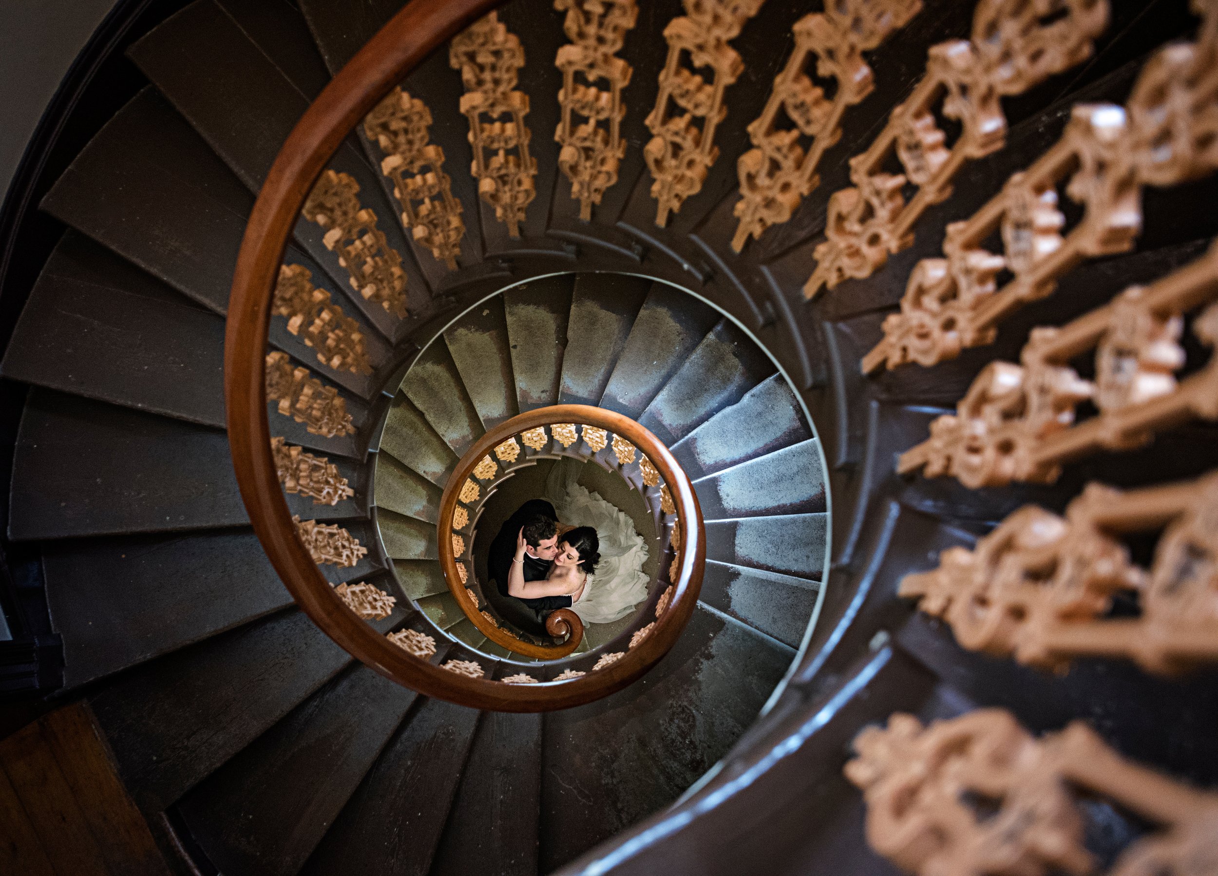 A couple in wedding attire embracing at the bottom of a spiral staircase, viewed from above.