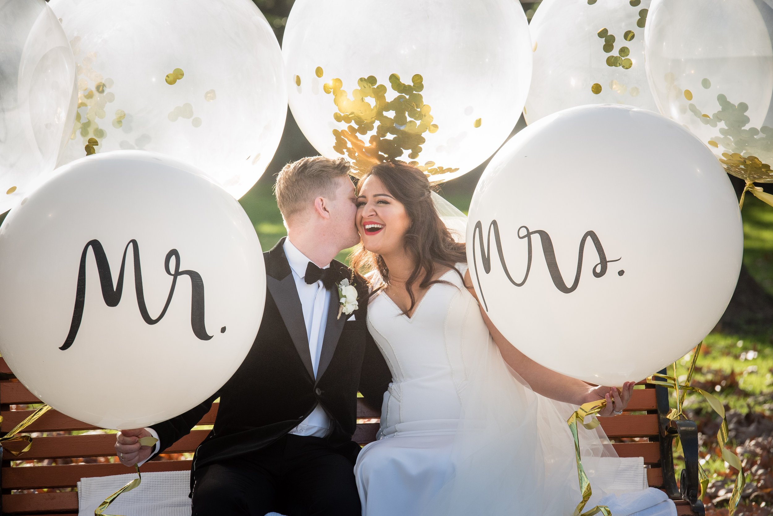 A bride and groom sit on a park bench surrounded by white balloons with 'Mr.' and 'Mrs.' written on them, celebrating their wedding with the groom kissing the bride on her cheek.