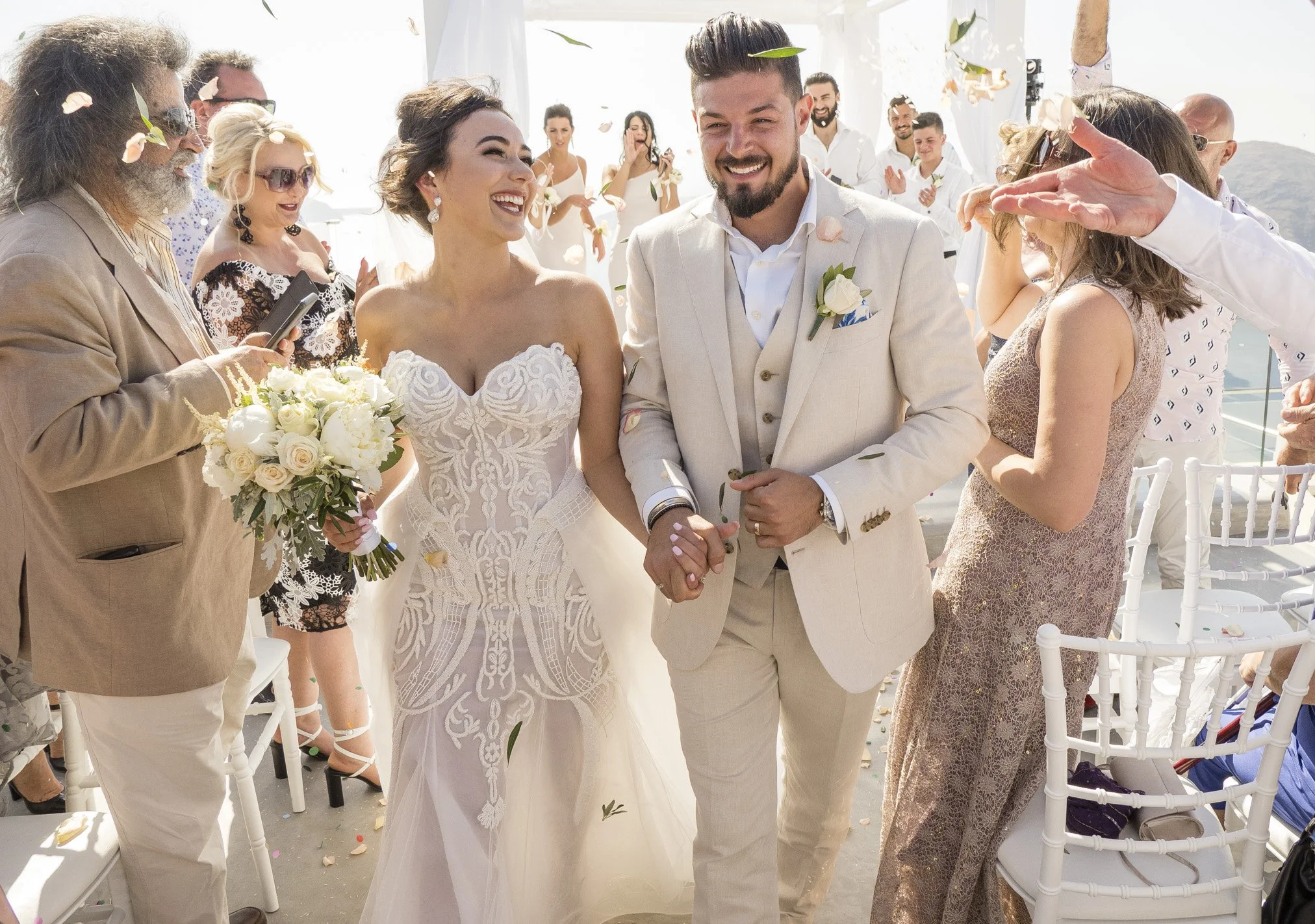 A smiling bride and groom holding hands during their outdoor wedding celebration, surrounded by guests throwing flower petals and applauding.