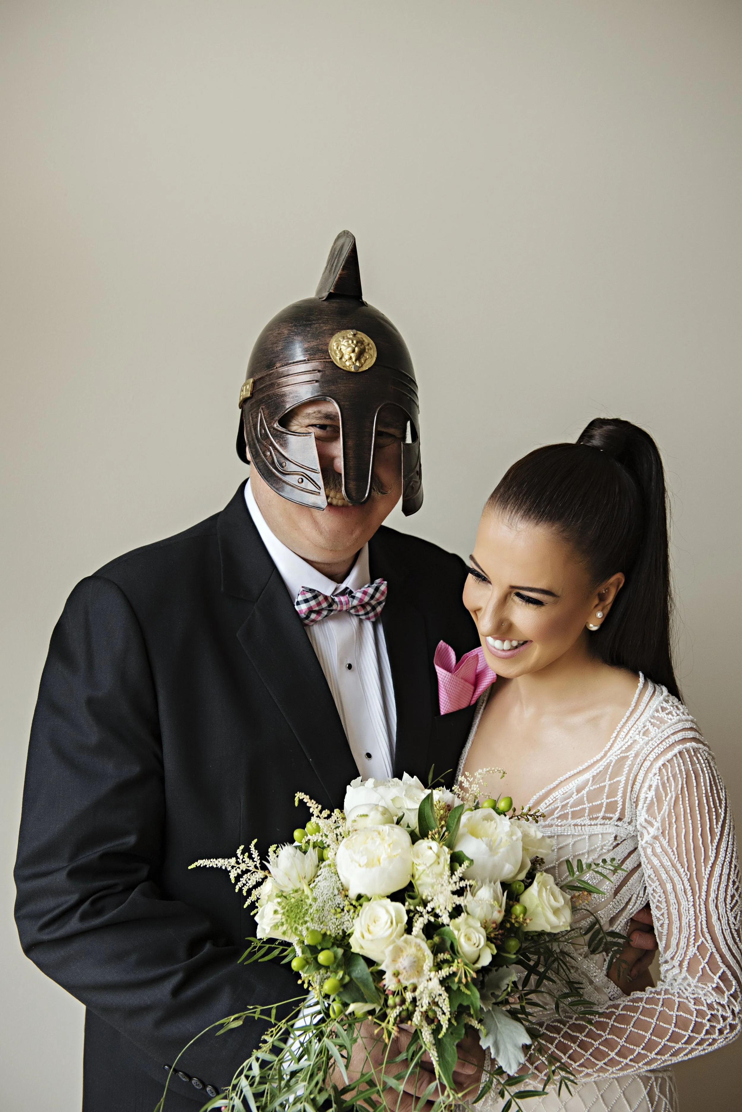 A bride and groom smiling, with the groom wearing a Roman helmet and a business suit, holding a bouquet of white flowers.