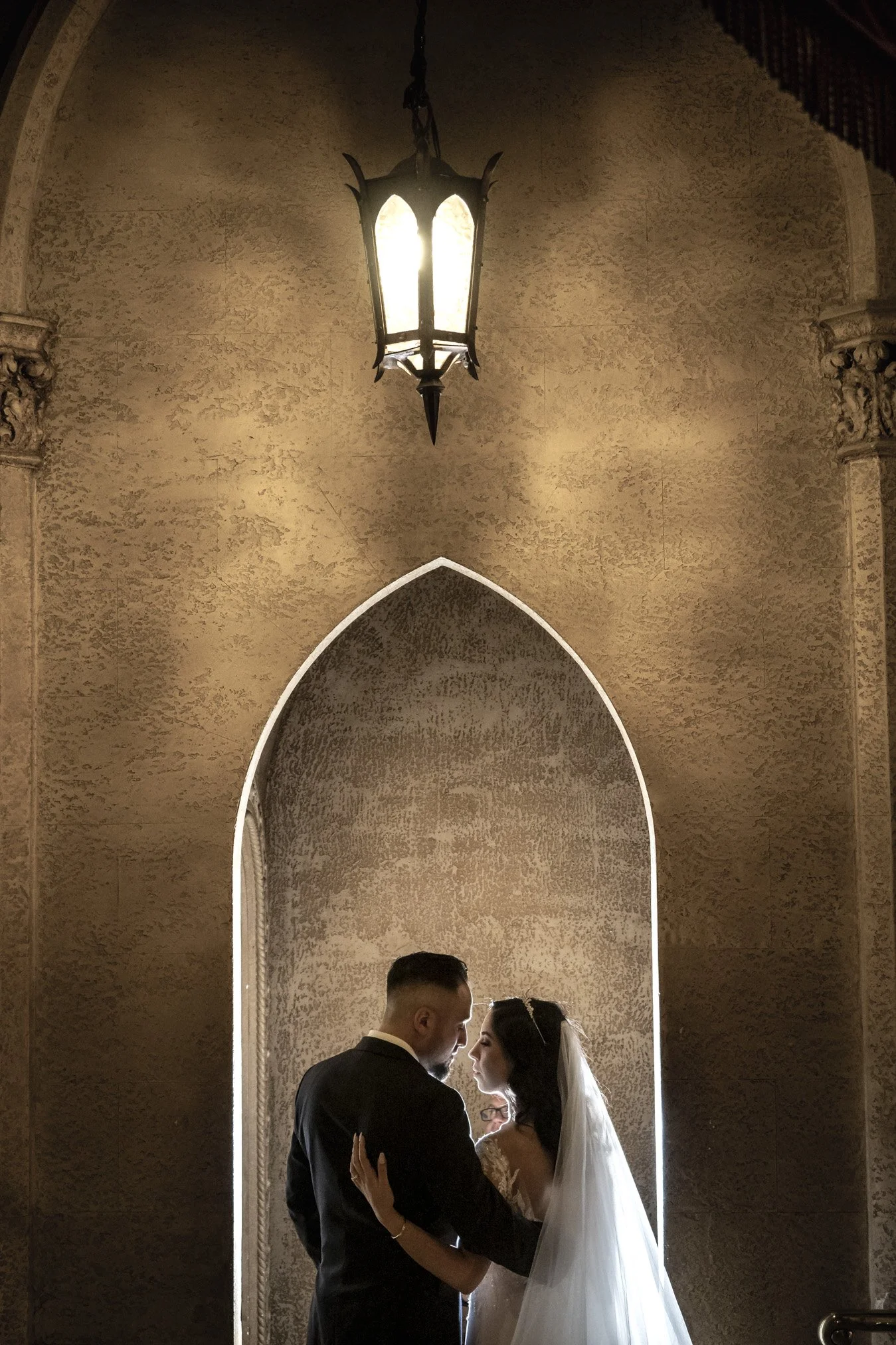 A bride and groom in a close embrace in front of an arched wall with a hanging lantern above, intimate moment during a wedding.