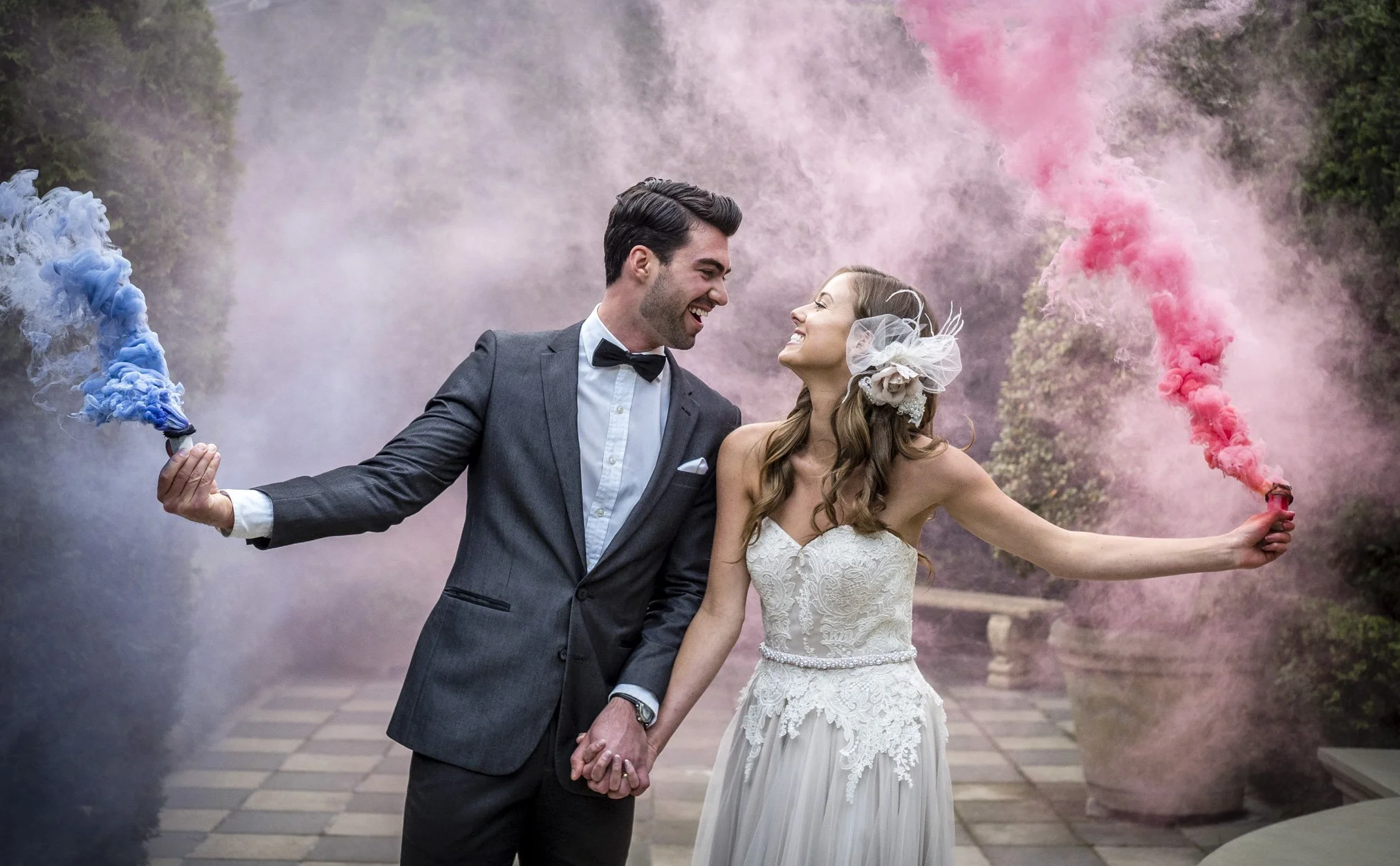 A newlywed couple holding hands and smiling at each other while holding colored smoke bombs, with pink and blue smoke in the air, outdoors on a checkered pathway surrounded by greenery.