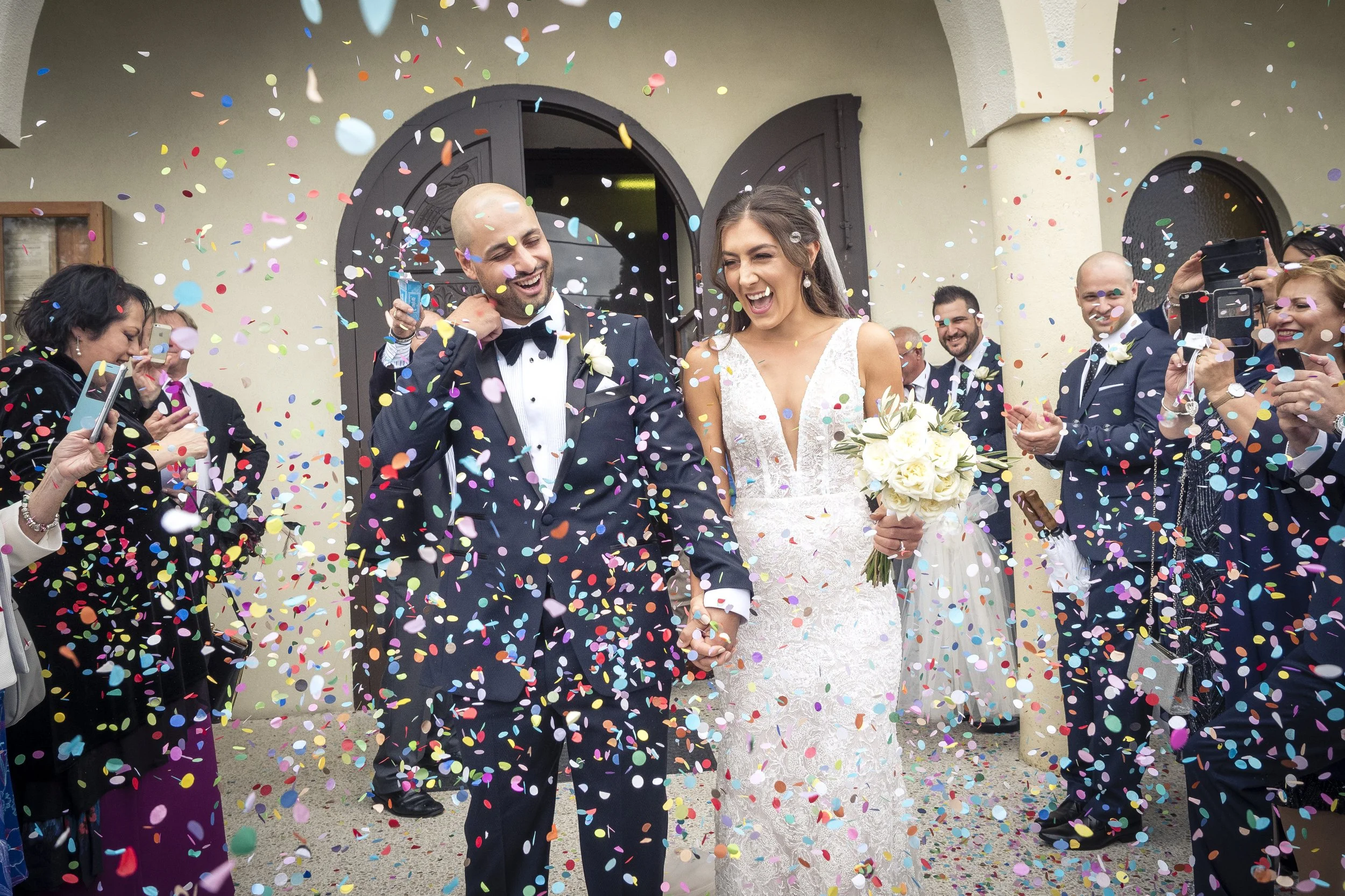 A bride and groom celebrating their wedding as colorful confetti falls around them, surrounded by friends and family taking pictures.