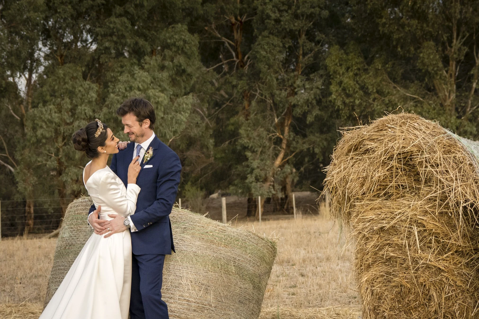 A bride and groom standing close together in a field with hay bales, trees in the background, during a wedding photoshoot.