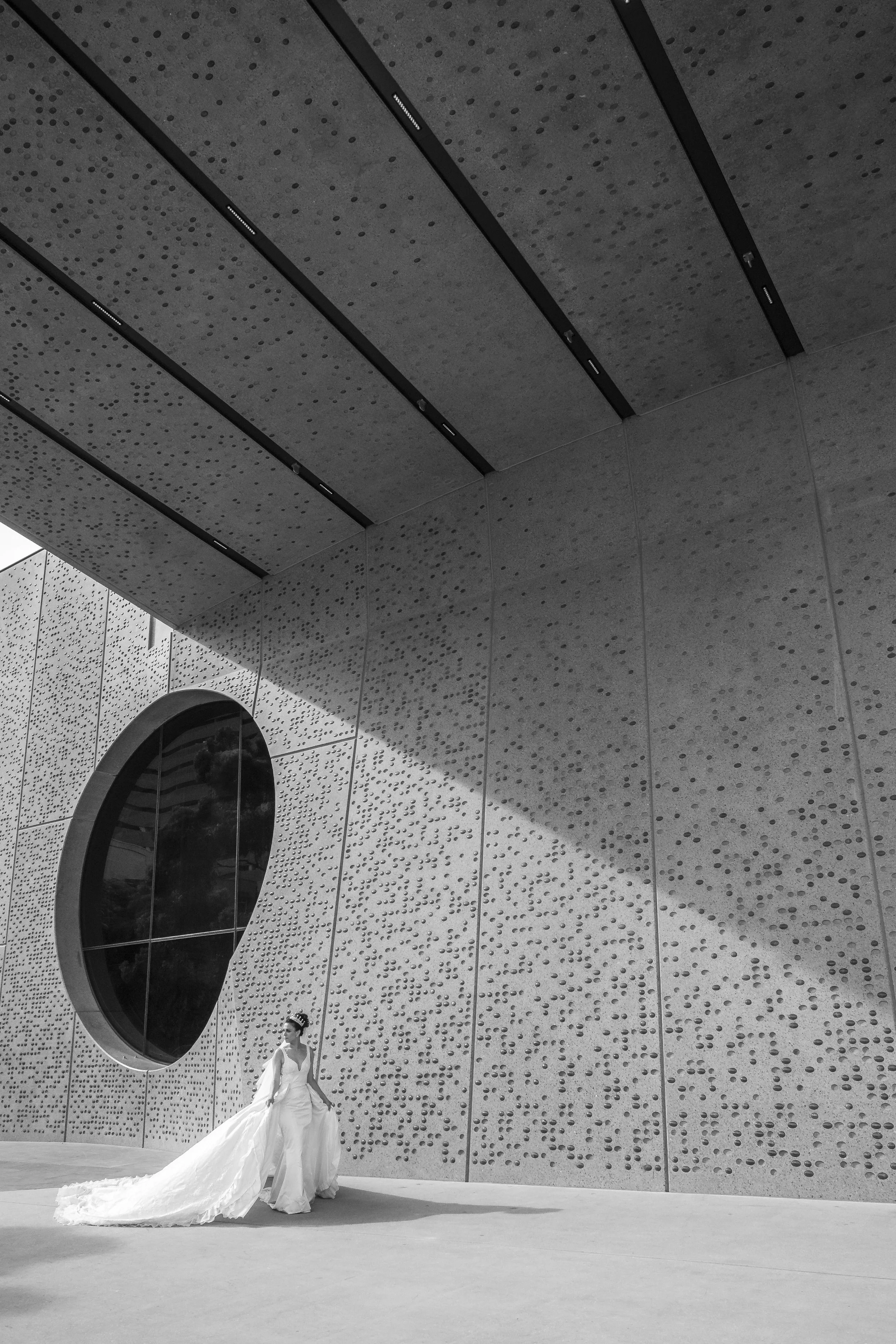 Black and white photo of a woman in a wedding dress standing outside a modern building with a textured facade and a large circular window.