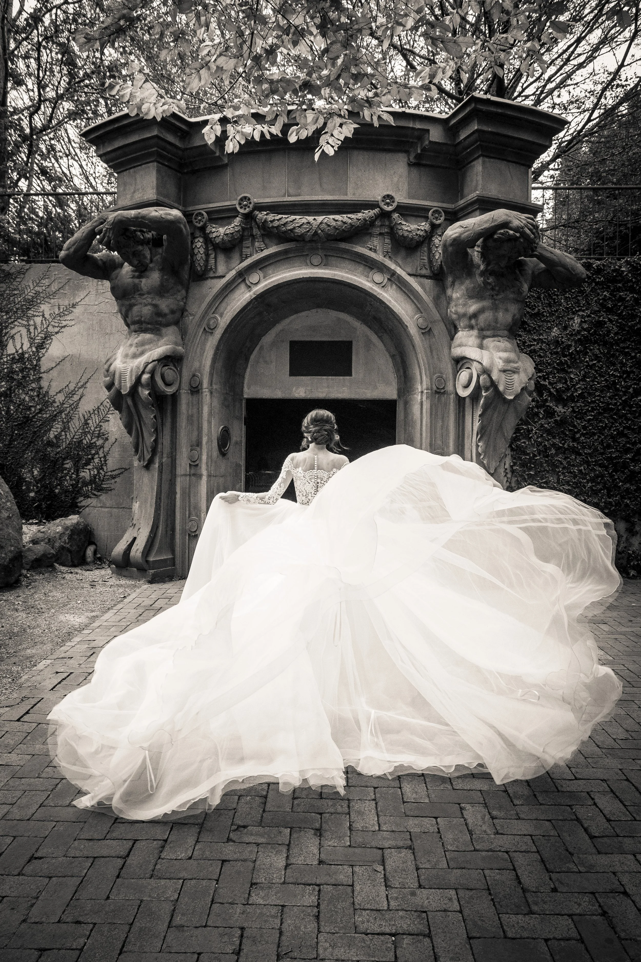 A bride in a wedding gown with a long, flowing skirt stands outdoors in front of a stone archway with ornate sculptures of two muscular figures on either side, surrounded by trees and foliage.