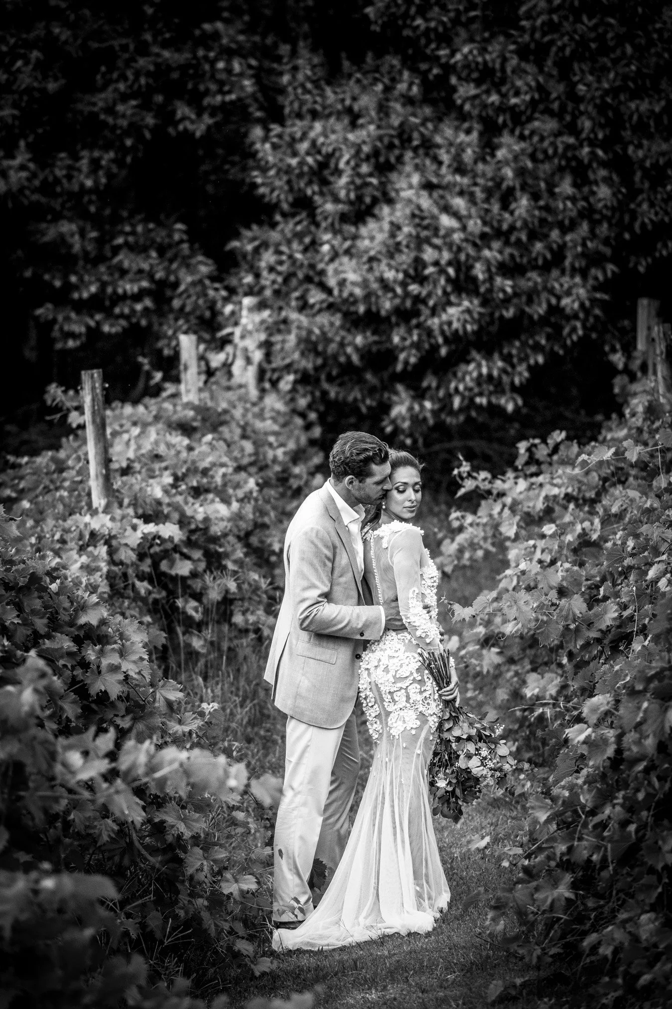 A black and white photograph of a couple in wedding attire standing in a vineyard, embracing each other with a woman holding a bouquet of flowers.