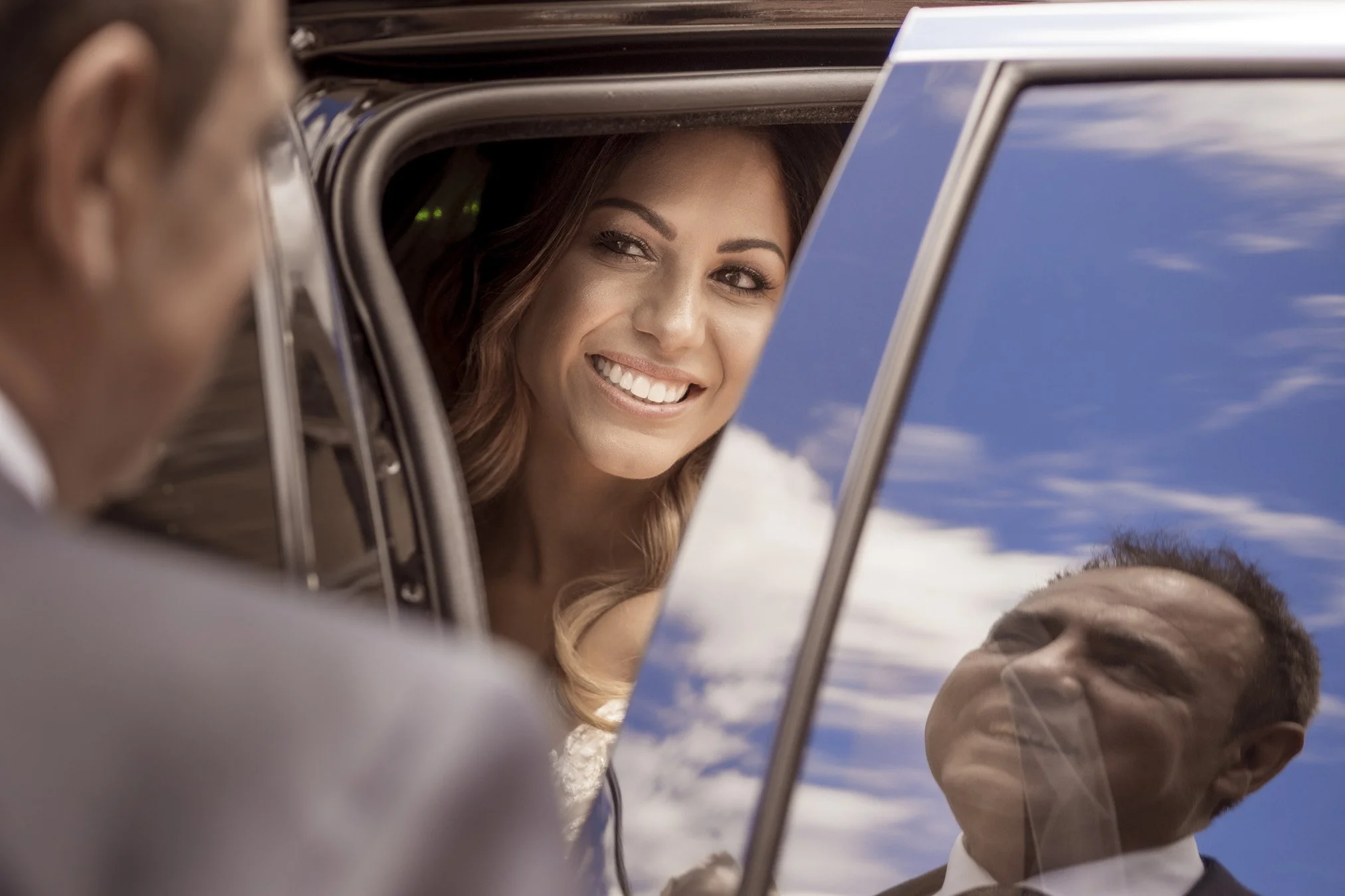 A woman smiling and looking out of a police car window at two officers, with a reflection of a blue sky and clouds on the car window.