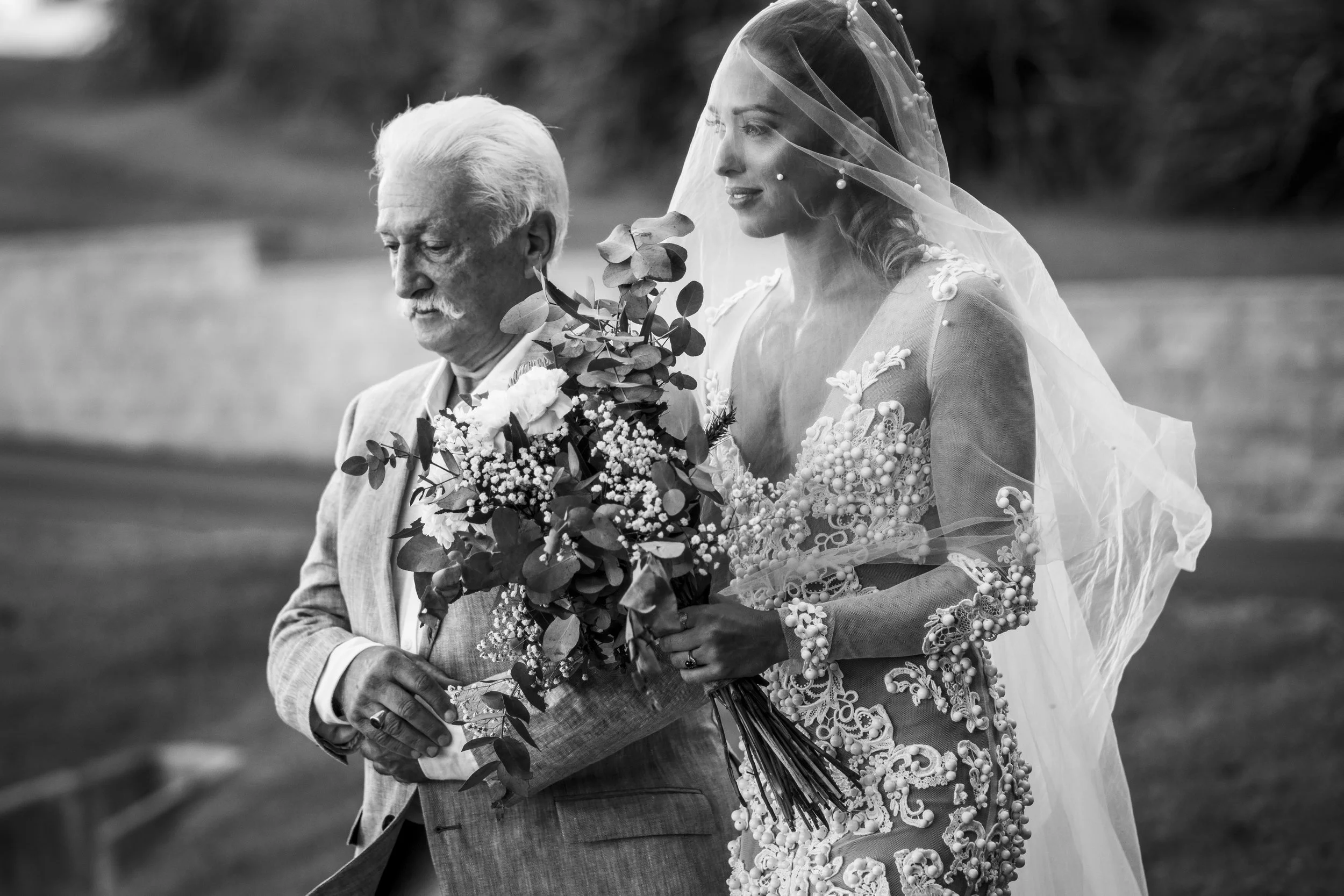 A bride in a wedding dress and veil holding a bouquet of flowers walking outdoors with an older man in a suit, in black and white.