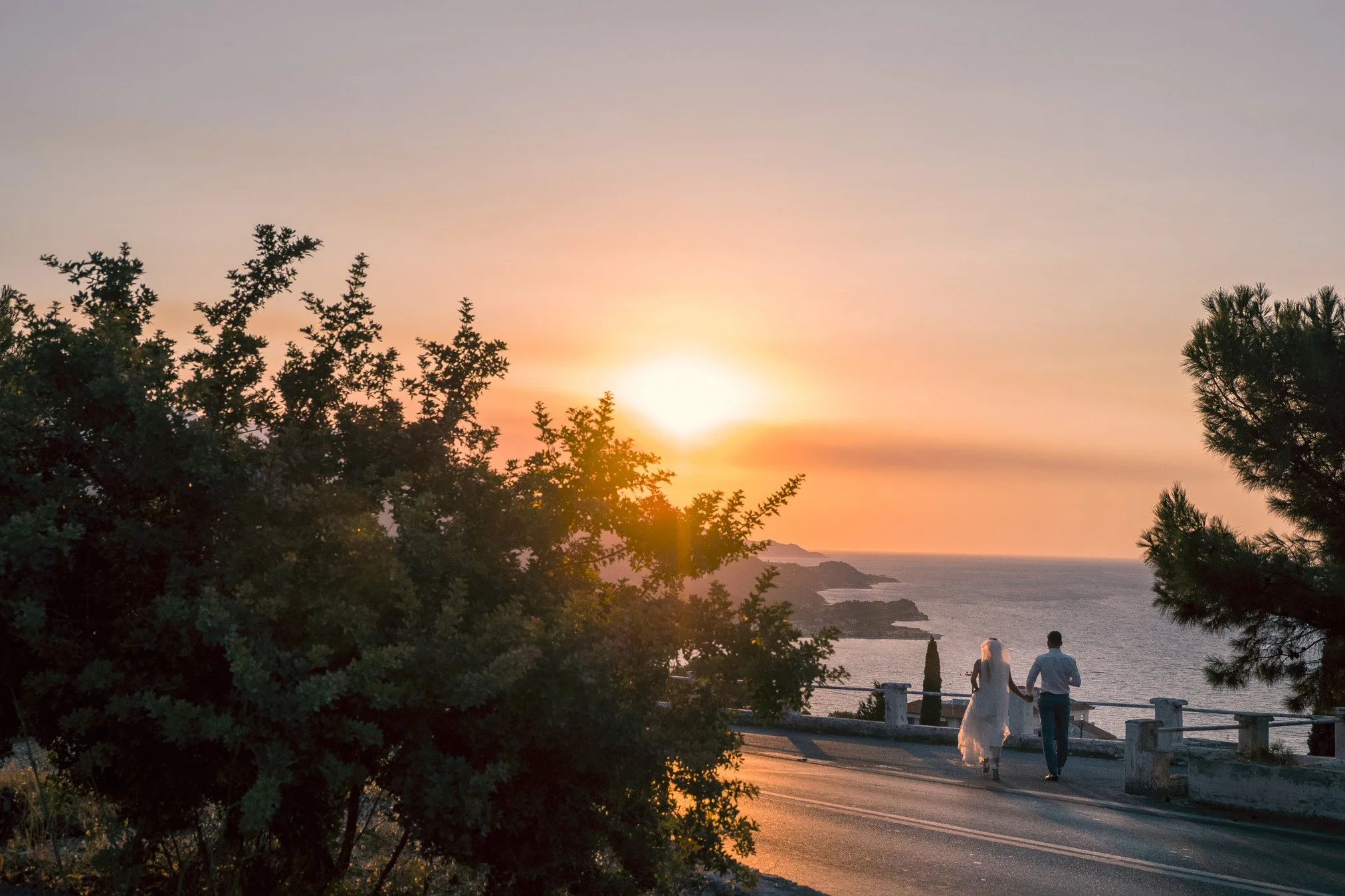 A couple walking hand in hand along a seaside road at sunset, with trees in the foreground and a scenic view of the ocean and distant islands.