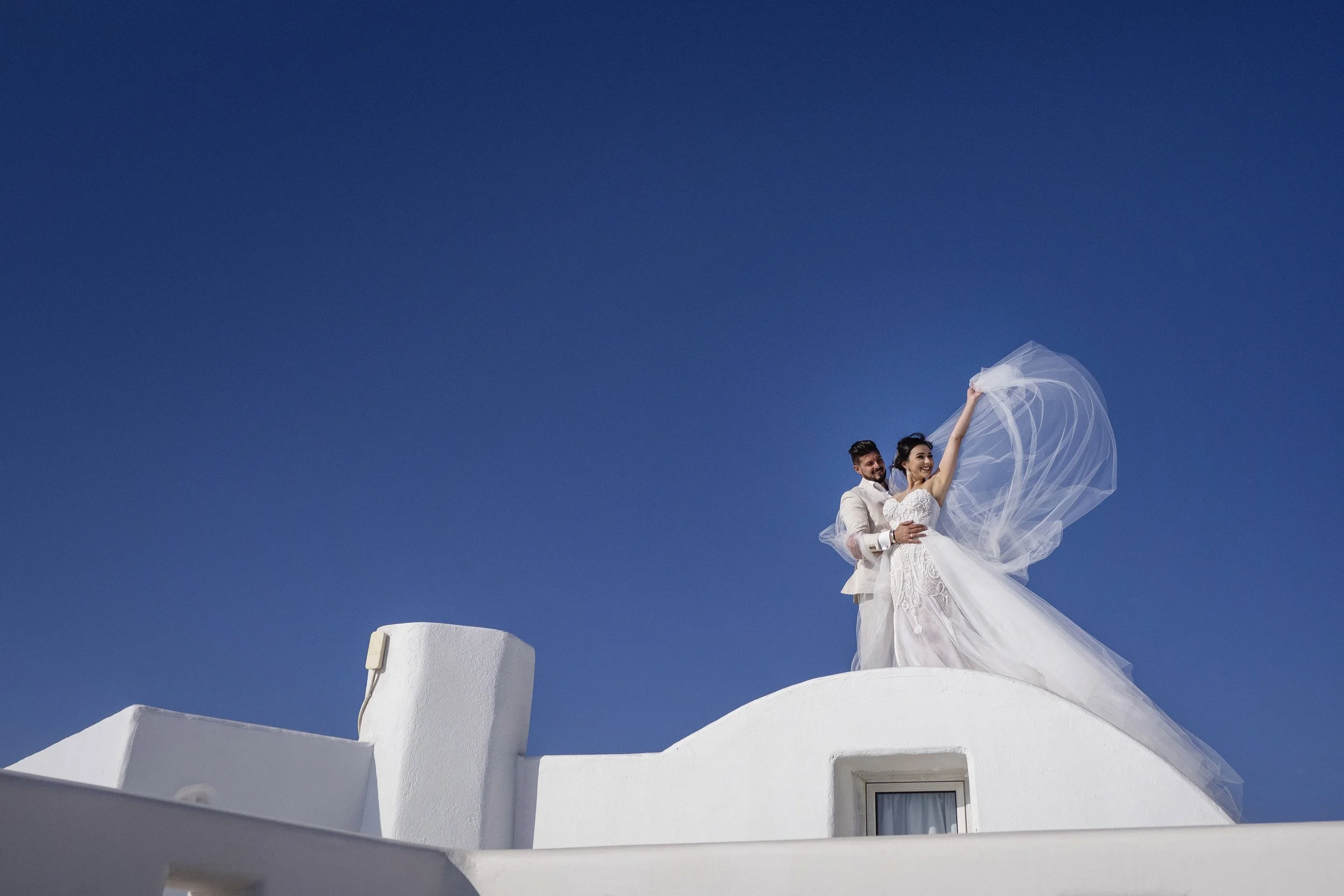 A bride and groom in wedding attire standing on a white rooftop with a clear blue sky background, celebrating their wedding with the bride holding her veil up.