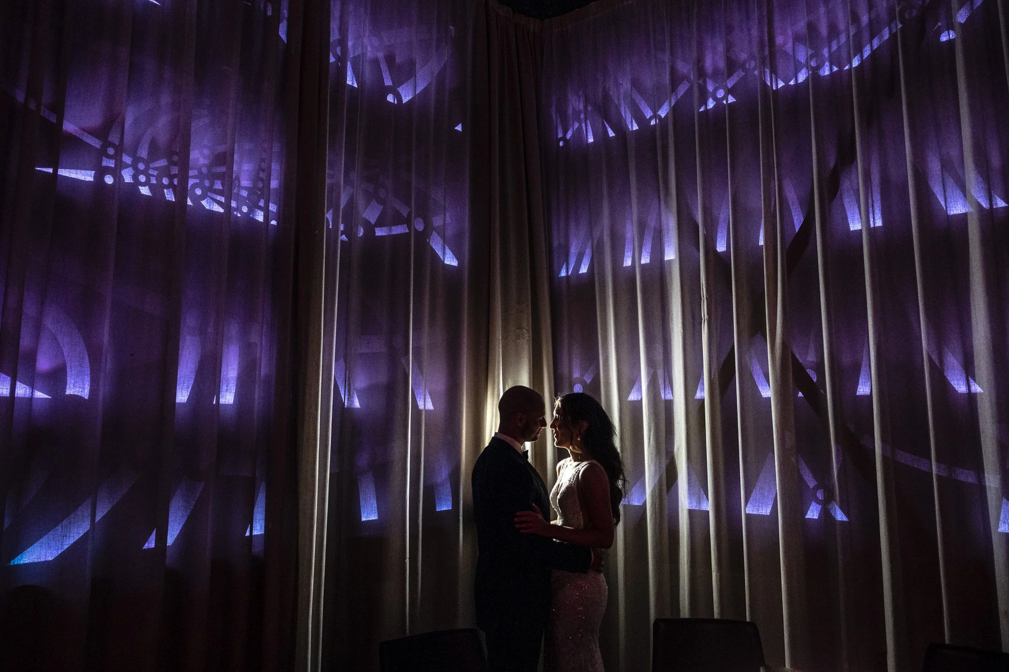 Silhouette of a couple in formal attire standing close together in front of a large curtain with abstract purple clock face projections.
