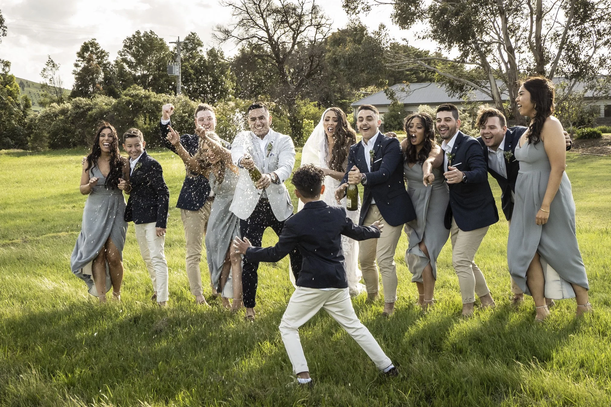 A group of people celebrating outdoors, spraying champagne and joyfully dancing on a grass field, with a young boy in the foreground.