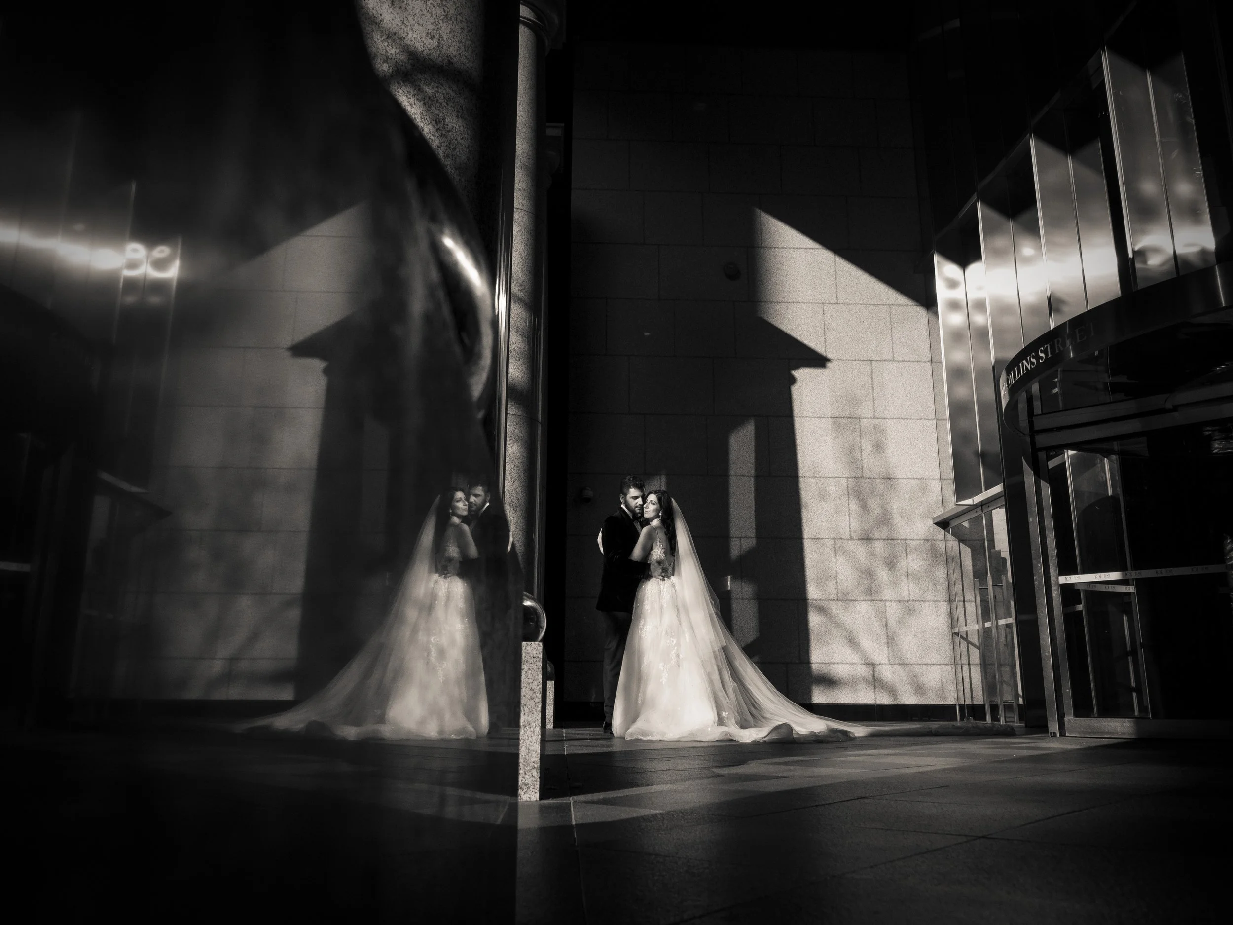 A black and white photo of a bride and groom standing close together, with their reflection visible in a shiny, curved glass surface on the left, in front of a modern building with large concrete walls and glass doors. The bride is in a wedding gown 