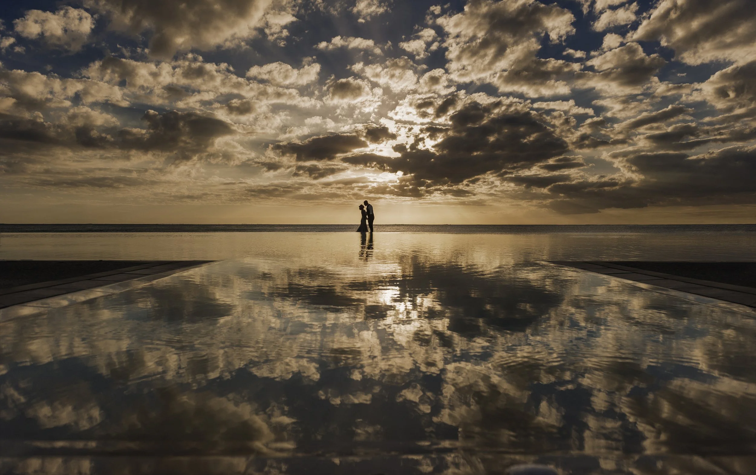 A silhouette of a couple standing close together on a beach at sunset, with a cloudy sky reflected on a nearby wet surface.