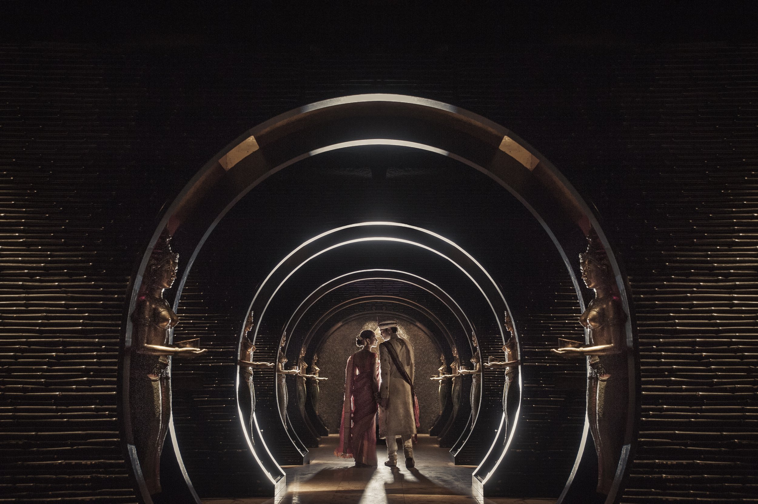 A couple walking through a dark, illuminated tunnel with circular arches and statue mannequins lining the sides.