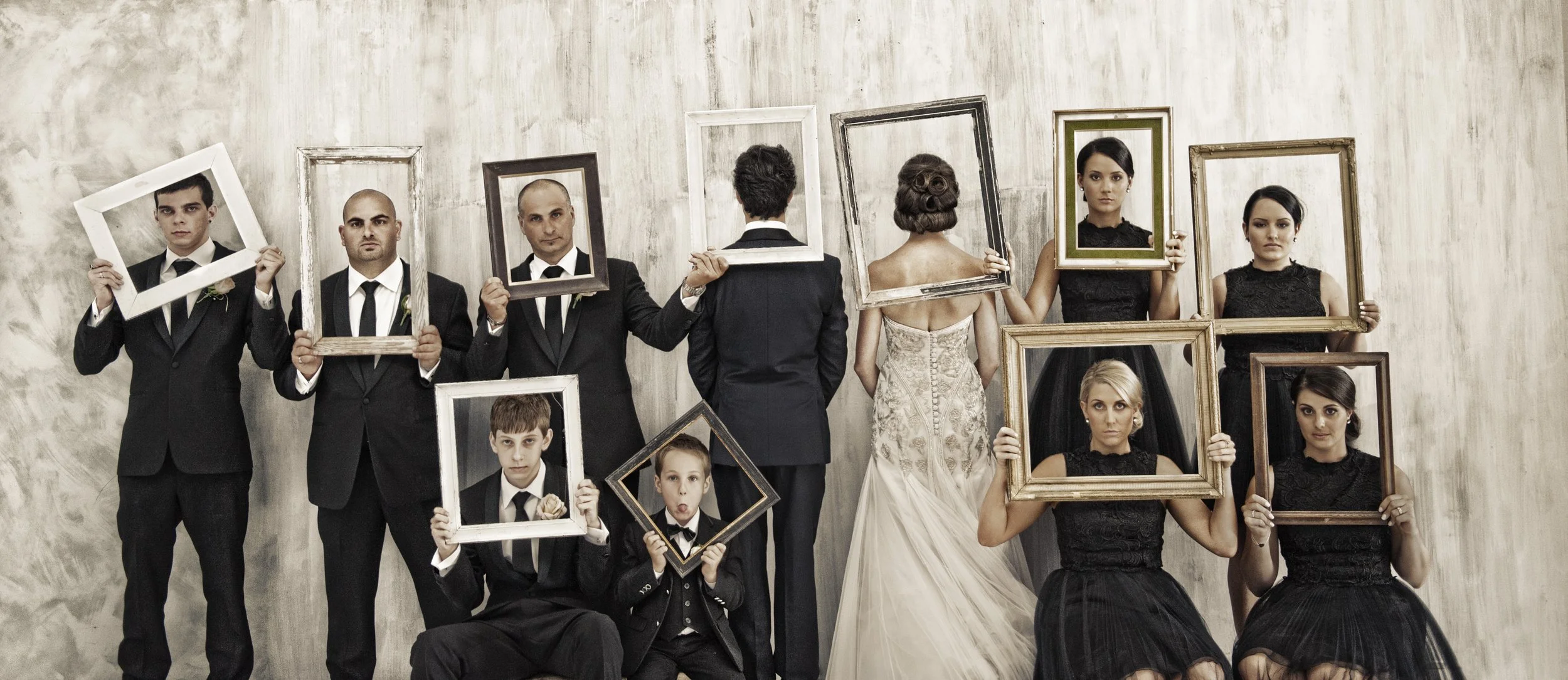 Group of wedding guests holding empty picture frames in front of a plain wall, with two women in bridal dresses and a man in a suit at the center.