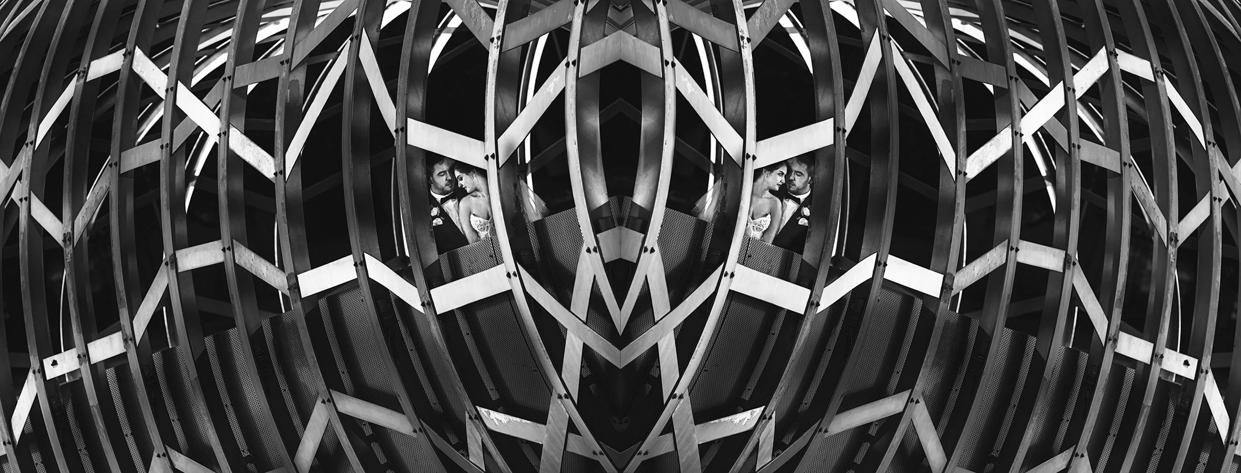 A black and white photo of a couple in wedding attire seen through the geometric framework of a modern, circular architectural structure with repeating curved metal beams.