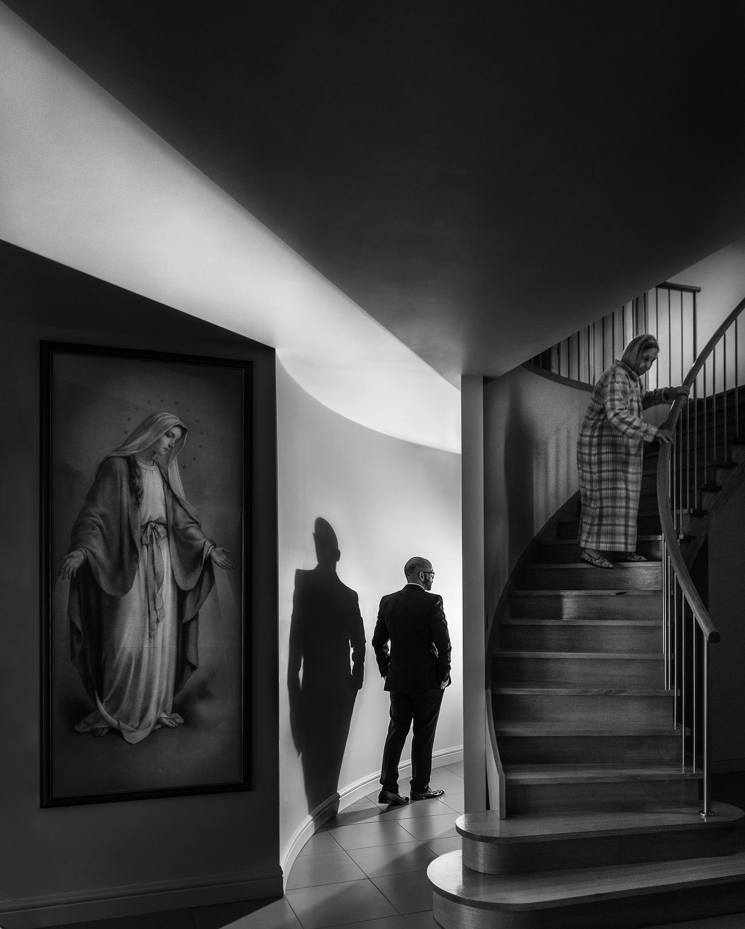 A black and white photo of two people in an art gallery, with religious artwork, including a portrait of the Virgin Mary. One person is ascending a curved staircase, and the other is standing on the floor, looking at the artwork.