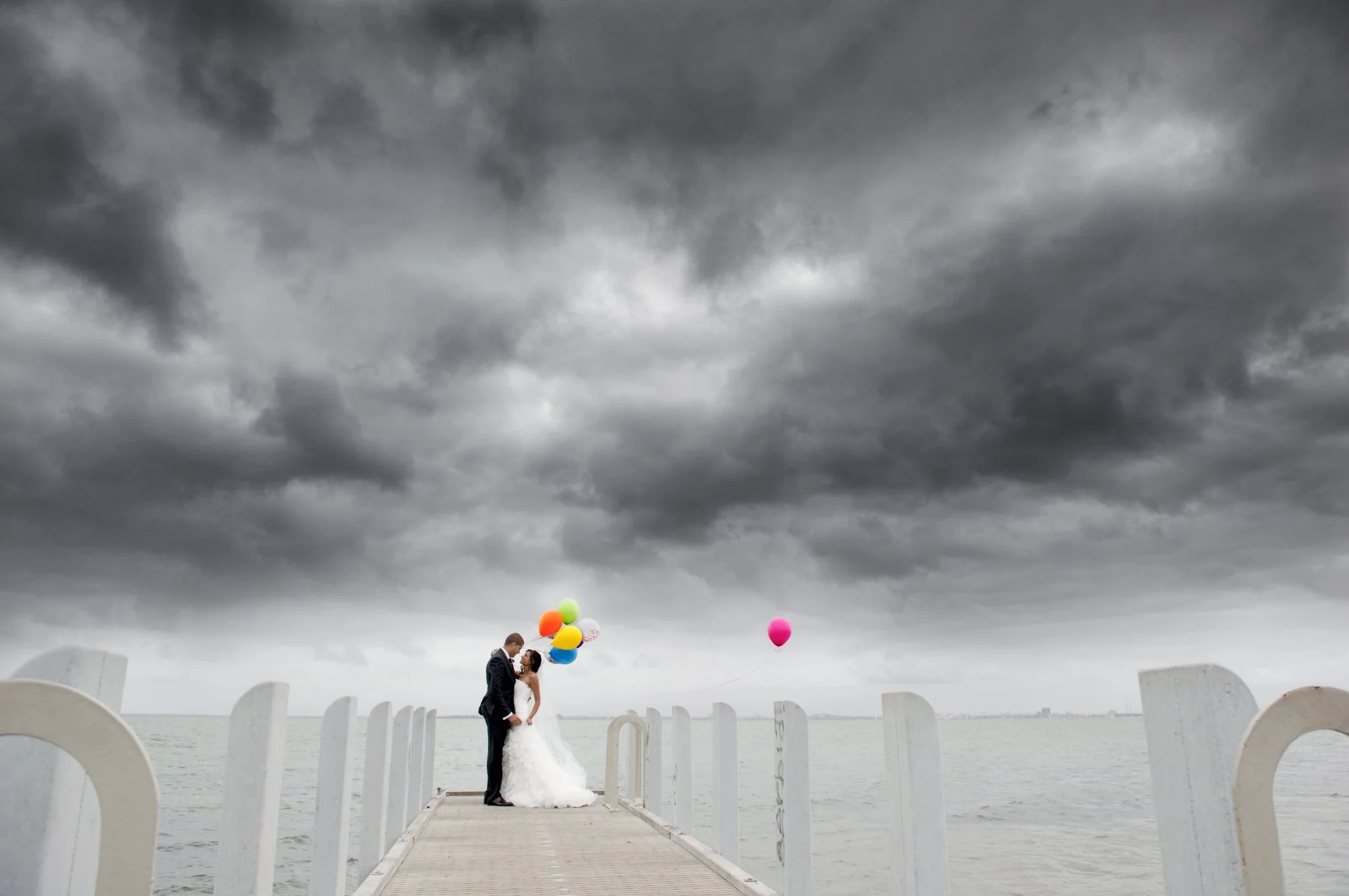 Bride and groom standing on a pier holding hands under dark, stormy skies, with colorful balloons floating behind them.