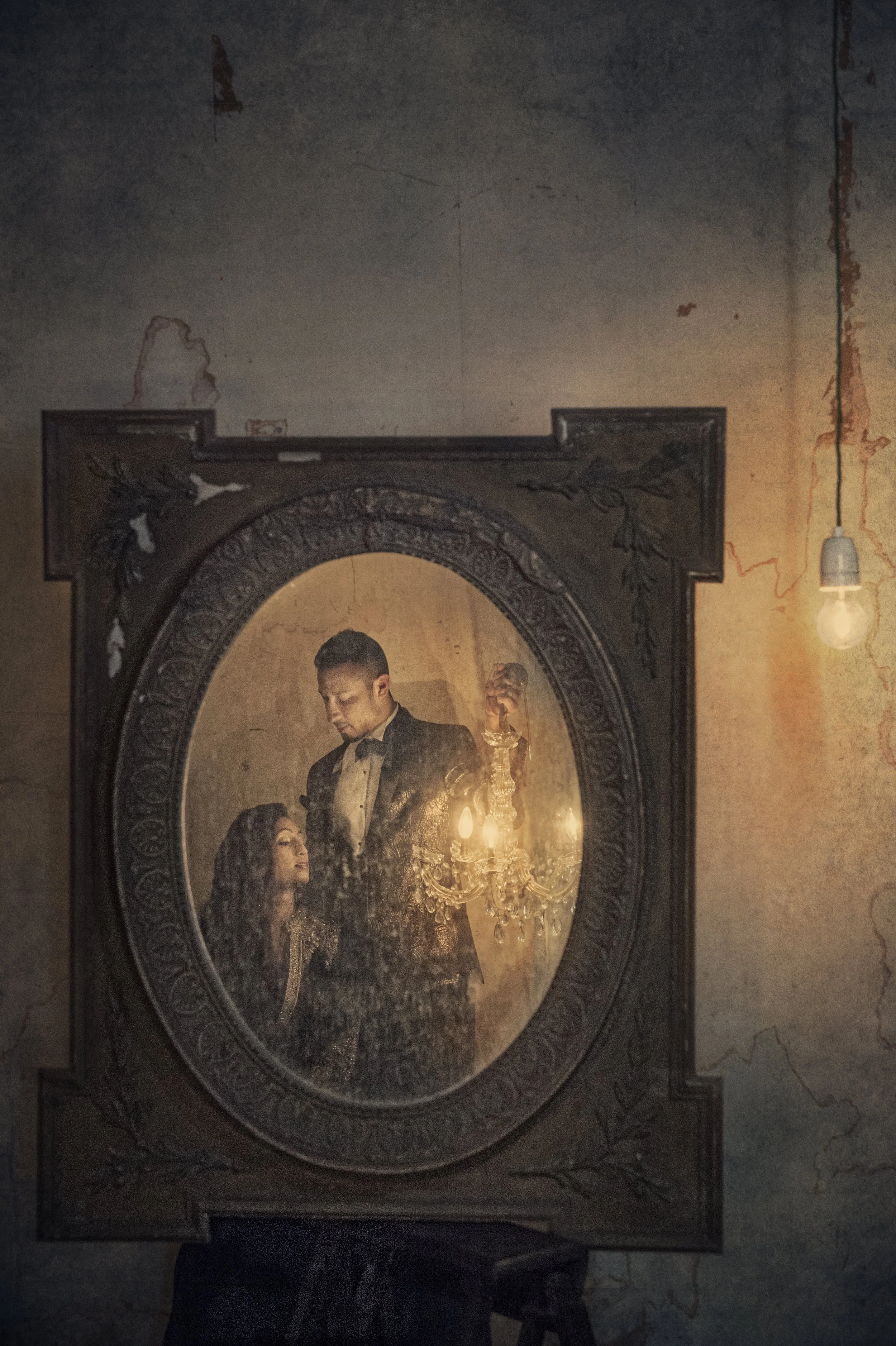A vintage framed mirror reflecting a man in a tuxedo and a woman in vintage attire, illuminated by a chandelier and a hanging light bulb, with a distressed wall background.