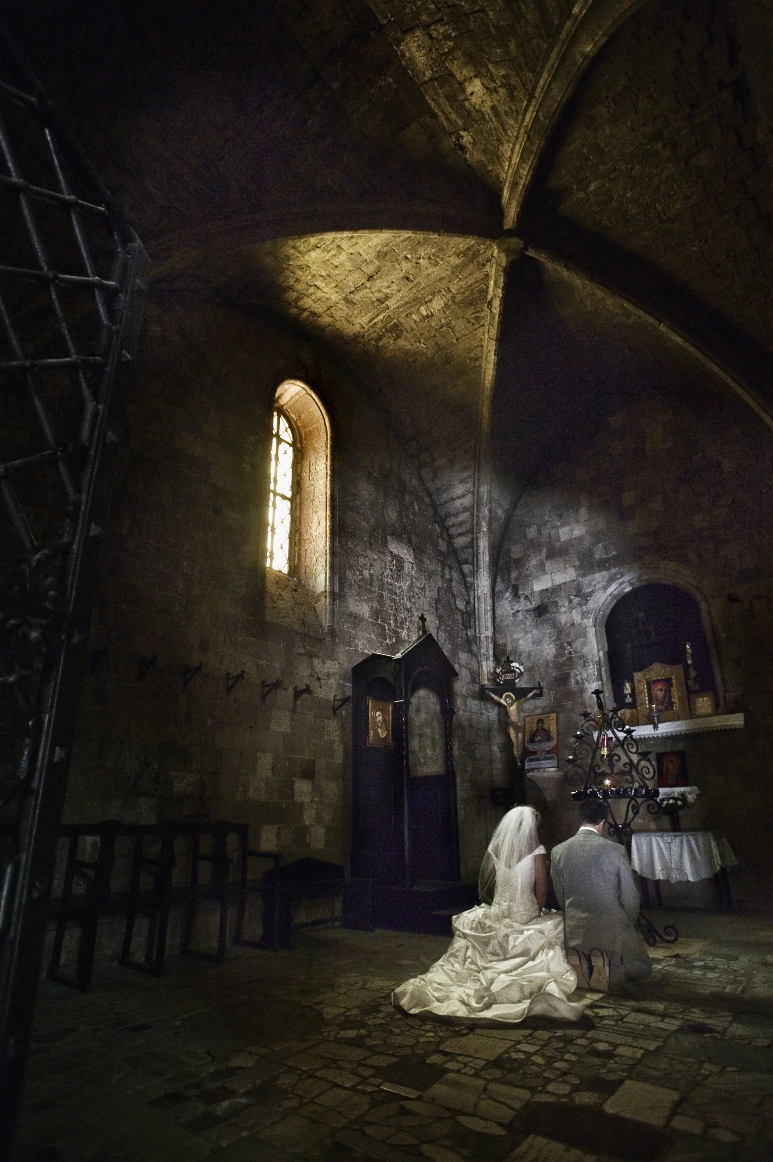 A bride and groom kneeling in a dimly lit stone church during their wedding ceremony, with religious icons, crucifix, and an altar in the background.