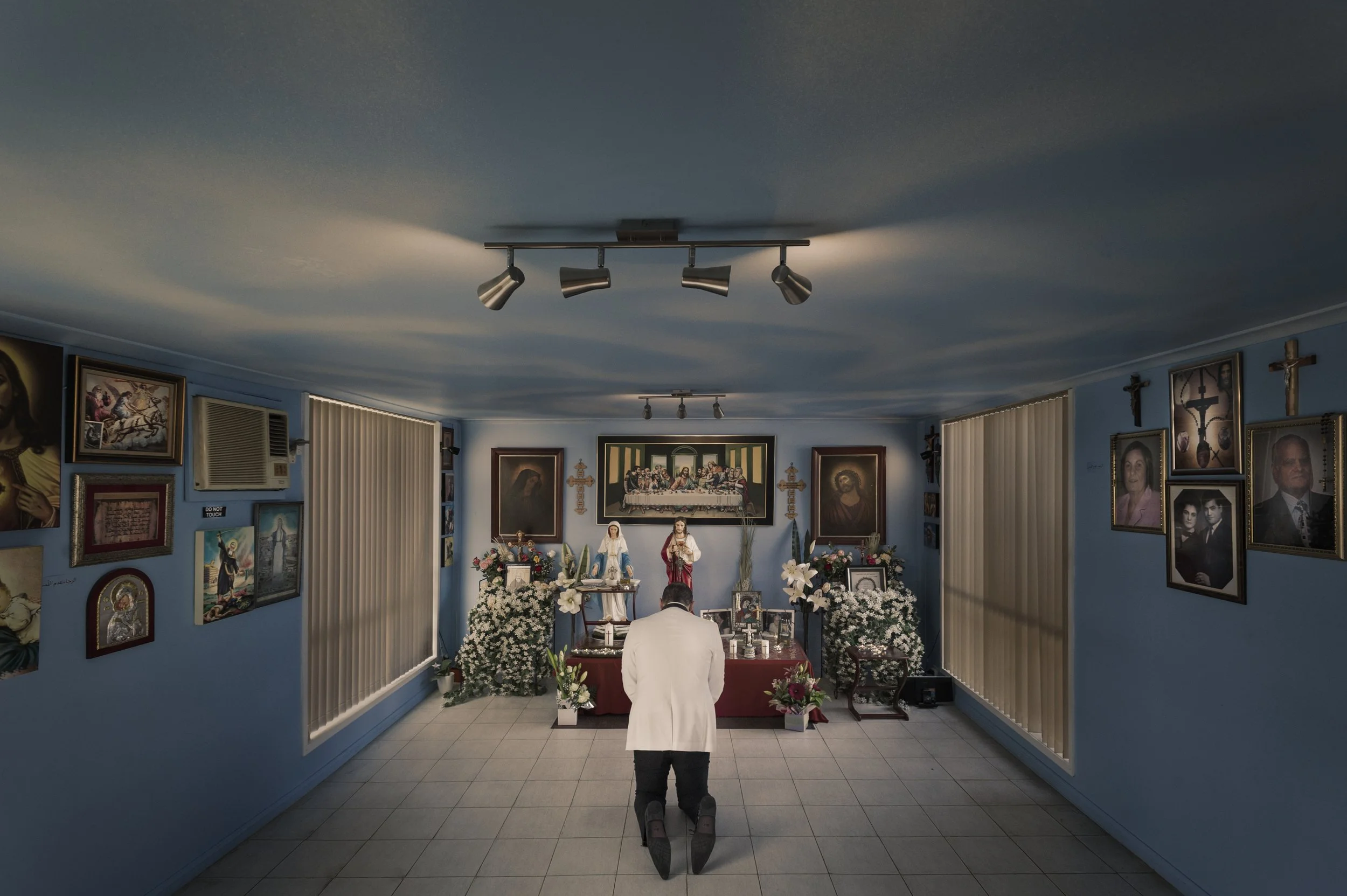 A person dressed in white kneeling before an altar with religious images and flowers in a small chapel.