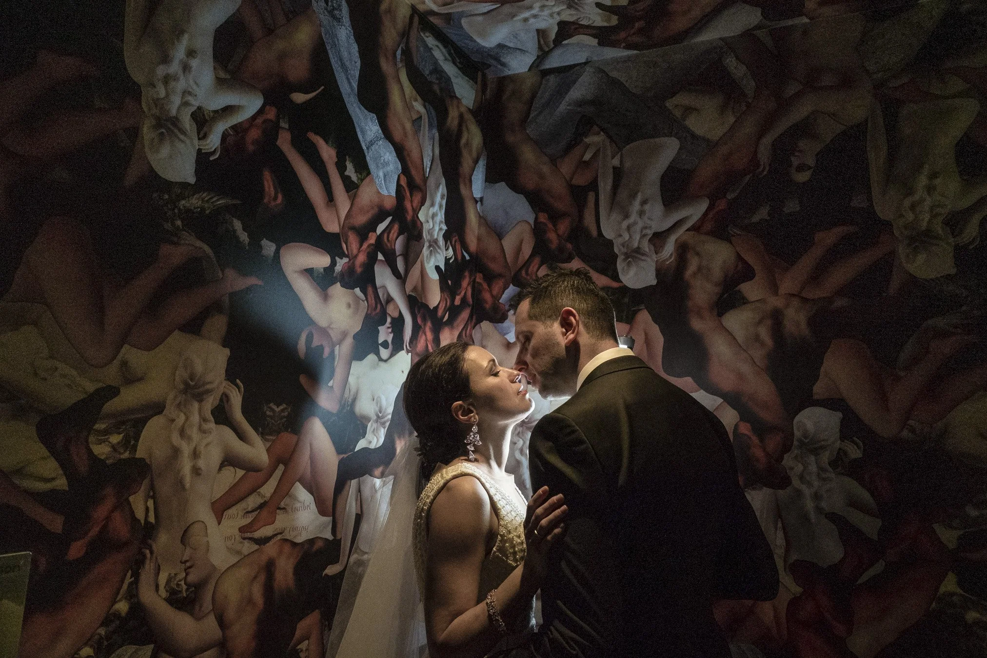 A bride and groom share a kiss in front of a wall adorned with classical art sculptures of nude figures.