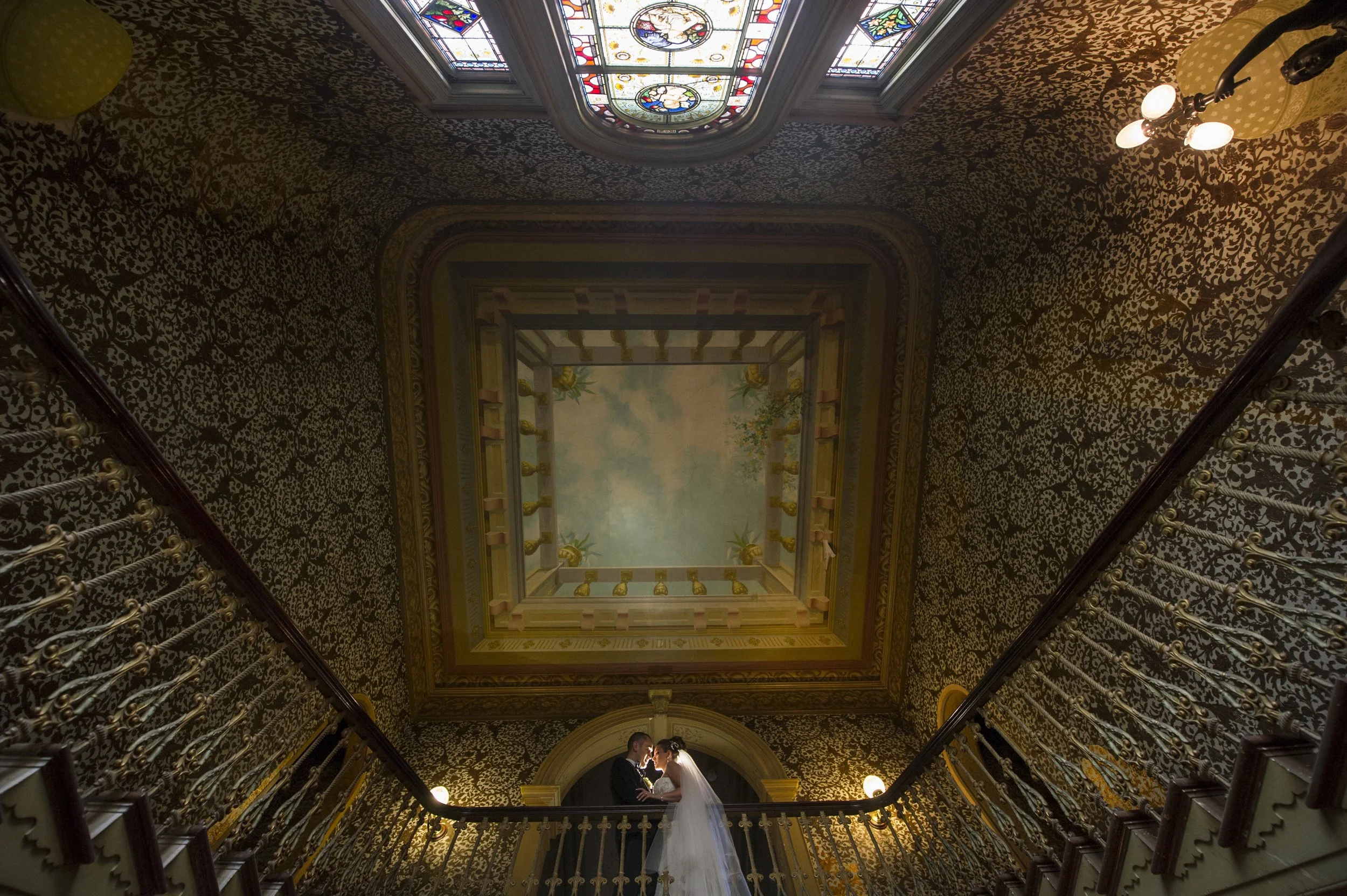 A bride and groom standing close together at the top of a staircase inside a grand, ornate building, with stained glass windows and decorative wallpaper on the ceiling.