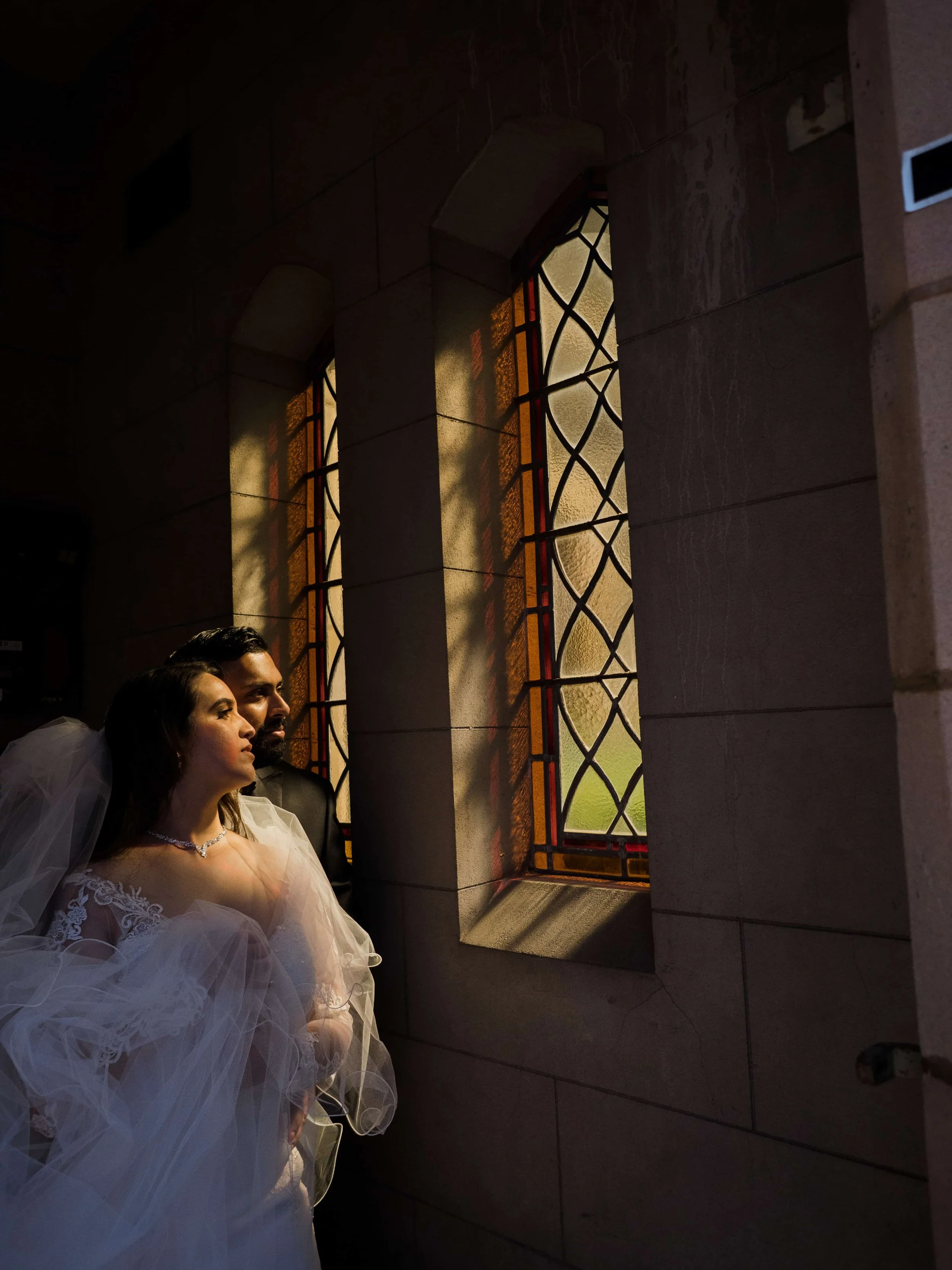 A bride and groom standing inside a building near a stained glass window, illuminated by natural light, with a contemplative look.