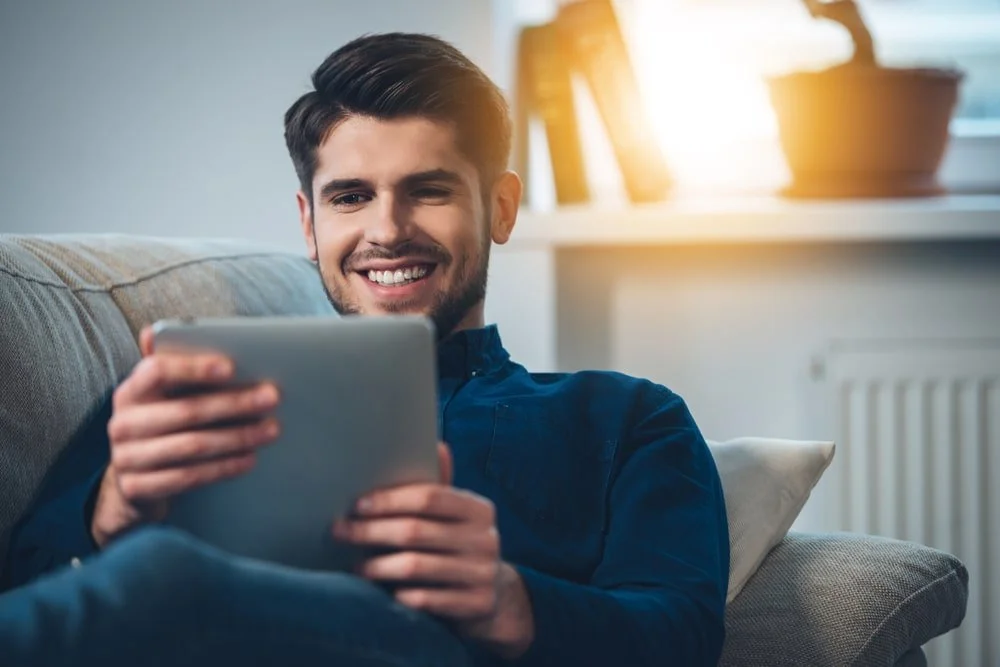 A young man with dark hair and a beard sitting on a sofa, smiling while looking at a tablet device. There is sunlight shining through a window in the background.