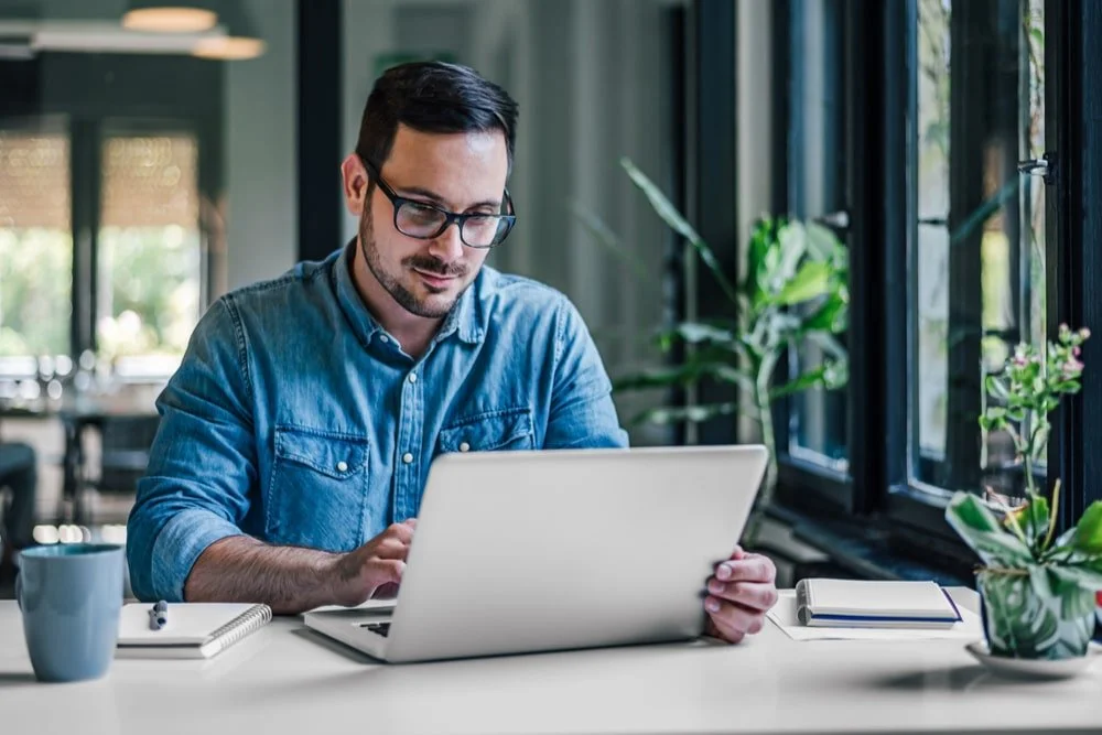 A man with glasses working on a laptop in a modern, well-lit office or cafe, with large windows and potted plants nearby.