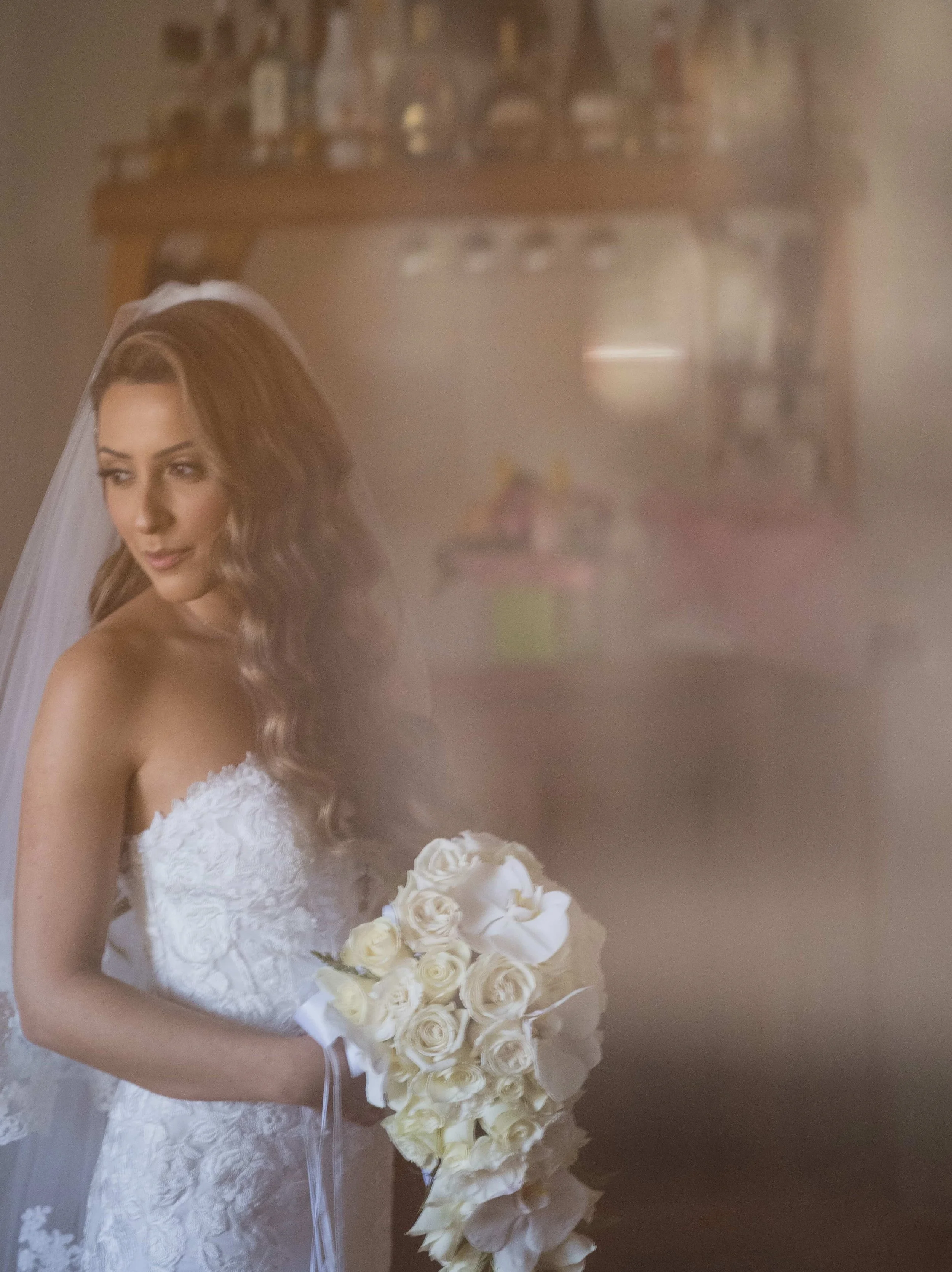 A bride with wavy brown hair wearing a white wedding dress and veil holding a bouquet of white roses, standing in a warmly lit room with a blurred background.