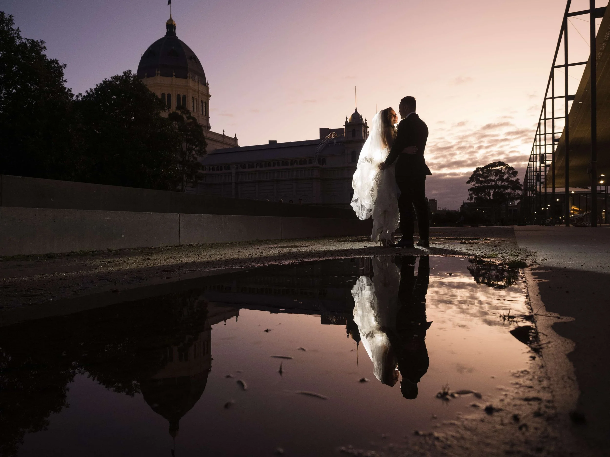 A bride and groom stand close together at sunset, with their reflection visible in a puddle on the ground and a domed historic building and trees in the background.
