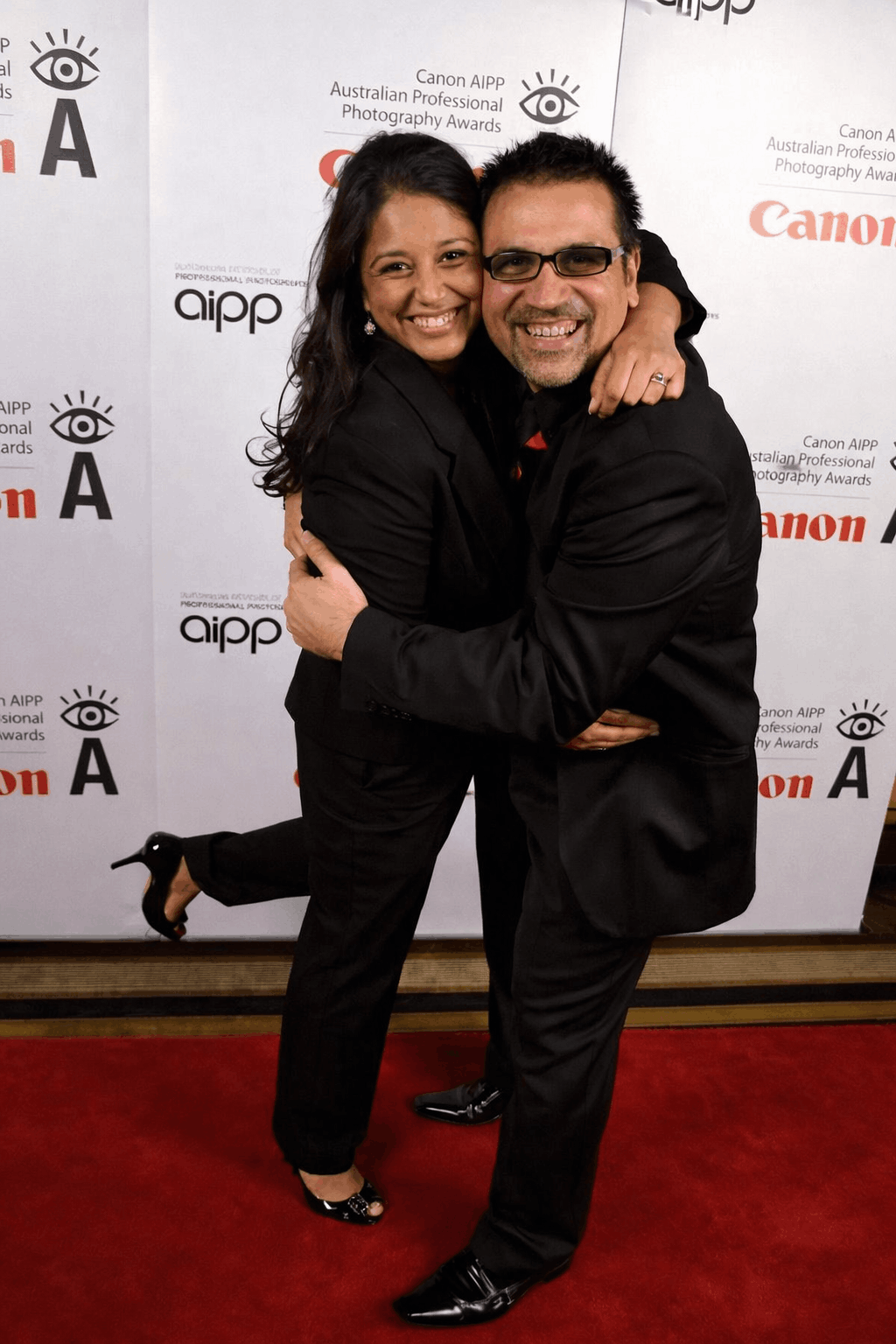 A man and woman hugging and smiling at a photography awards event, standing on a red carpet in front of a step and repeat banner with logos for Canon and AIPP.