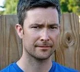 A man with short dark hair and a beard standing outdoors in front of a wooden fence.