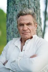 Older man with gray hair and a serious expression, standing outdoors next to a tree, wearing a white shirt.