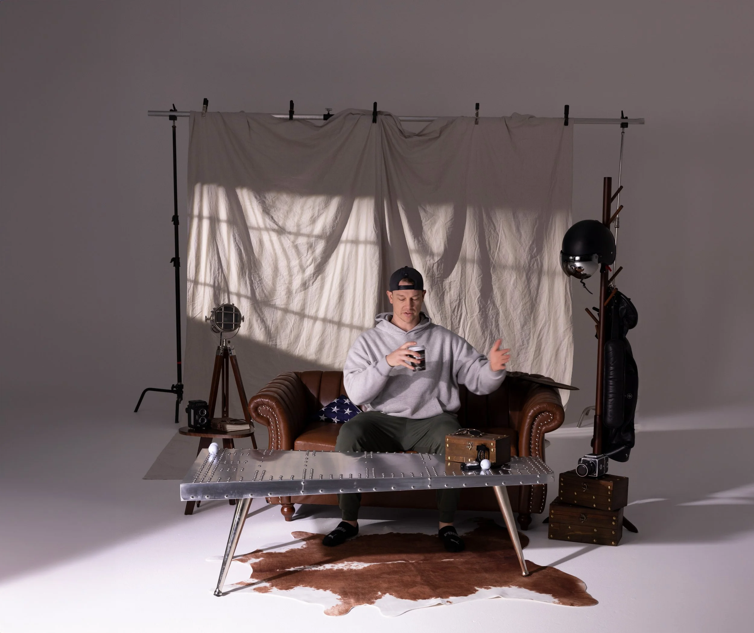A man in a gray hoodie sitting on a brown leather sofa looking at a smartphone. Behind him, there is a backdrop with a white sheet hanging from a metal support. On the left, a spotlight and a vintage film camera are set up. On the right, a metal coat rack with helmets and bags, and a small wooden chest are visible. A metallic table with rivets and a leopard print rug are in front of the sofa.