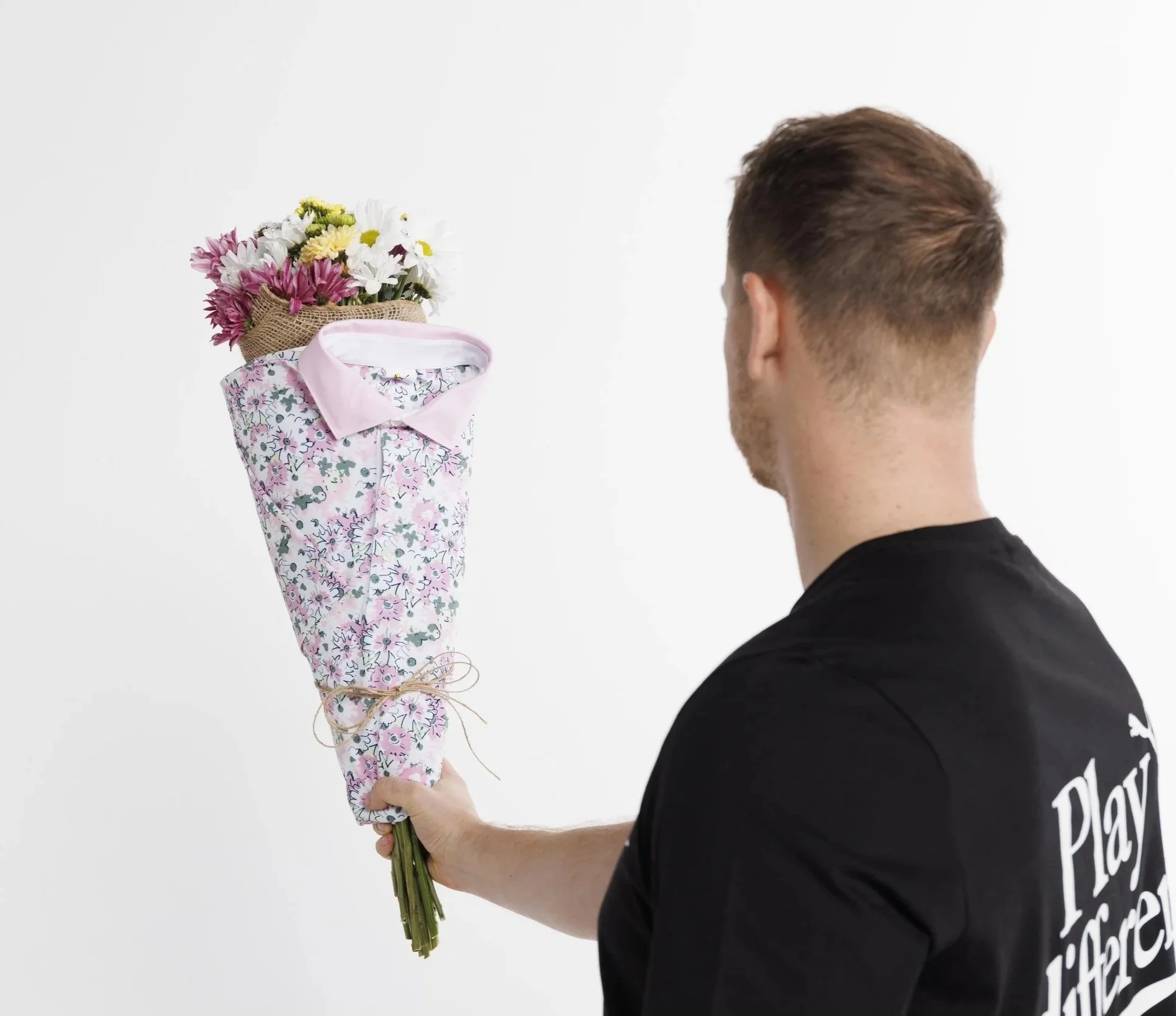 A person with short brown hair holding a bouquet of mixed flowers wrapped in floral paper and tied with string, against a plain white background.