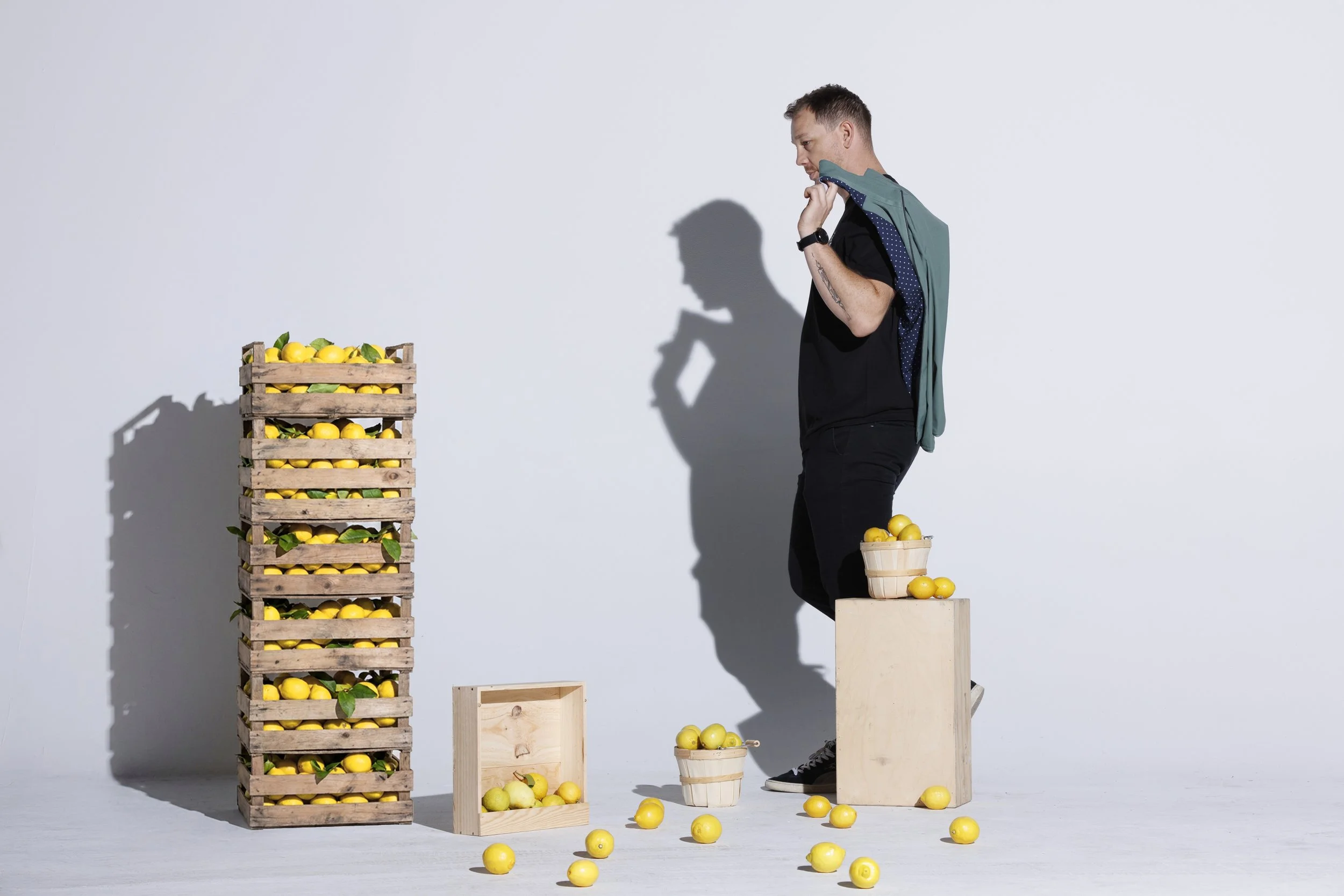 A man with a backpack holding a lemon in front of a stack of wooden crates filled with lemons, with some lemons spilled on the ground and a small wooden box.