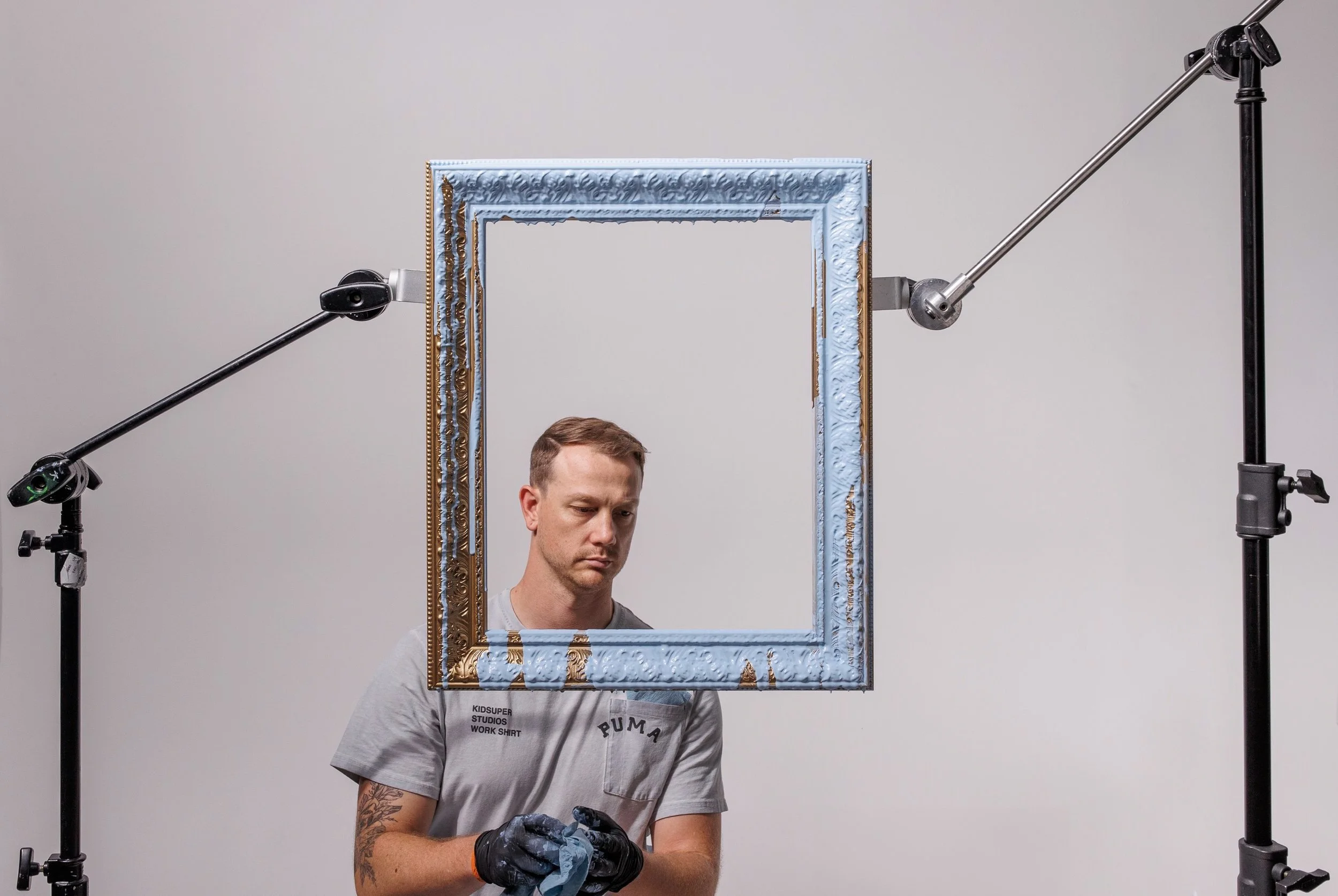 A young man with tattoos and wearing a gray T-shirt stands in front of a decorative framed mirror, surrounded by photo equipment, in a studio setting.