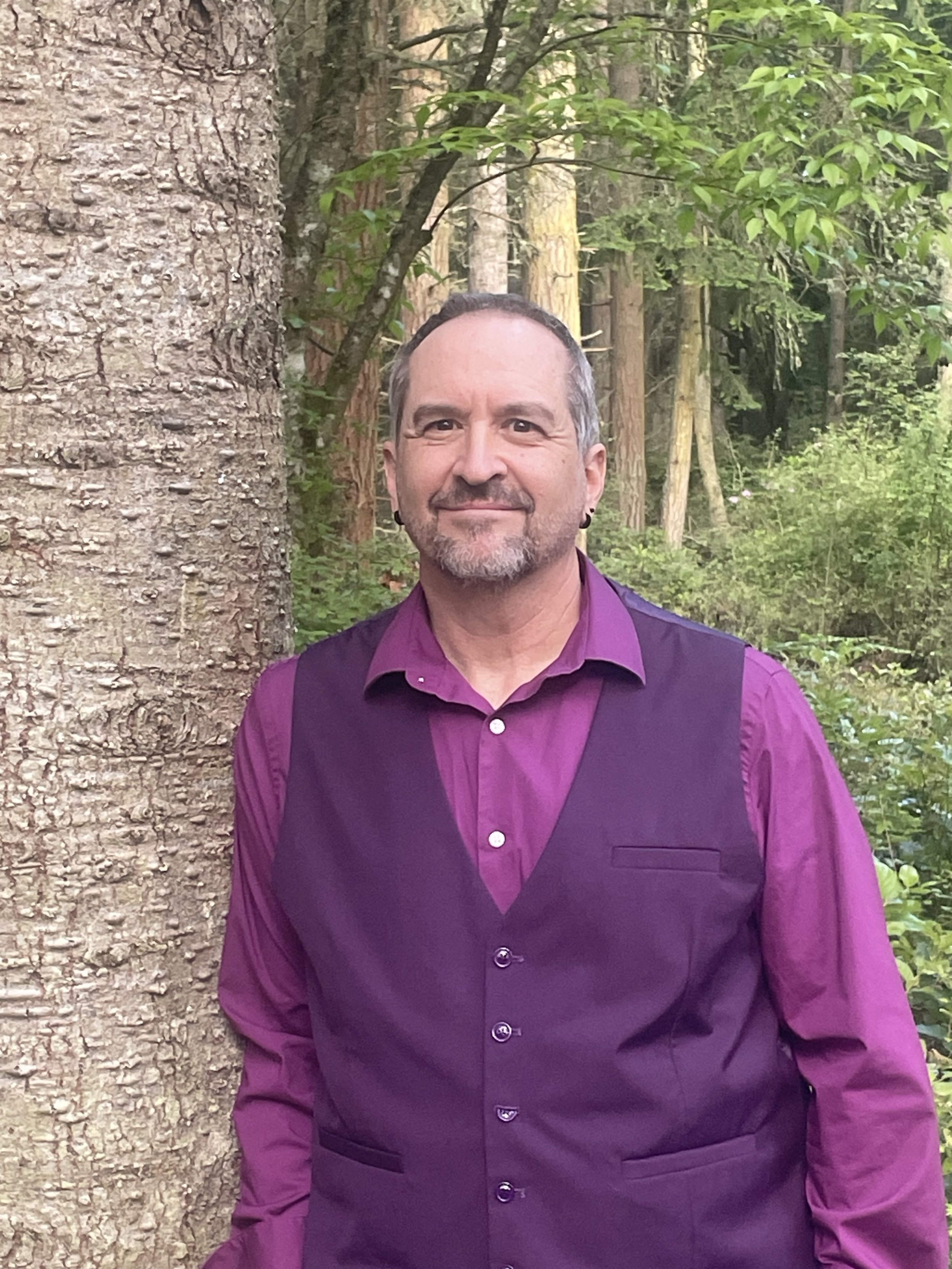 A man with short gray hair and a beard, wearing a purple shirt and matching vest, standing outdoors in a wooded area with trees and greenery.