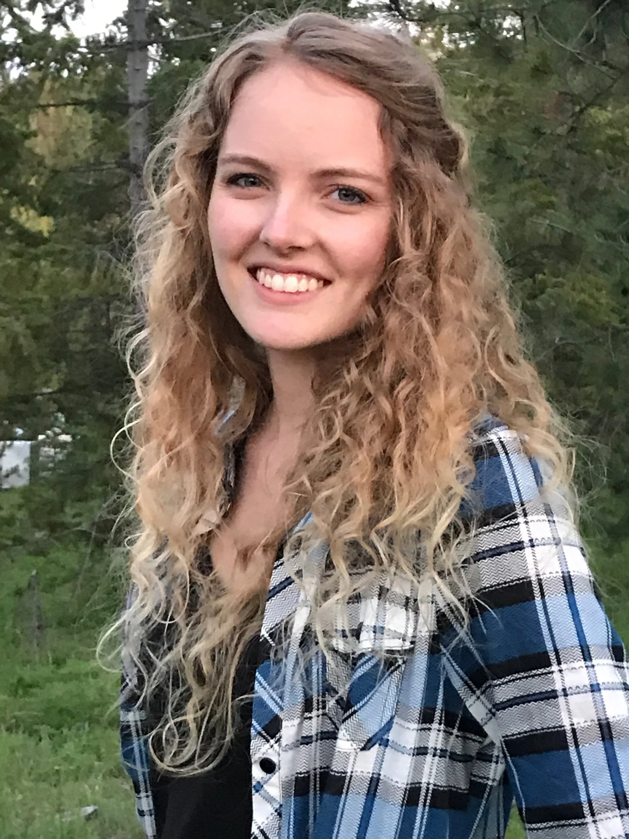 A young woman with long blonde curly hair smiling outdoors with trees in the background.