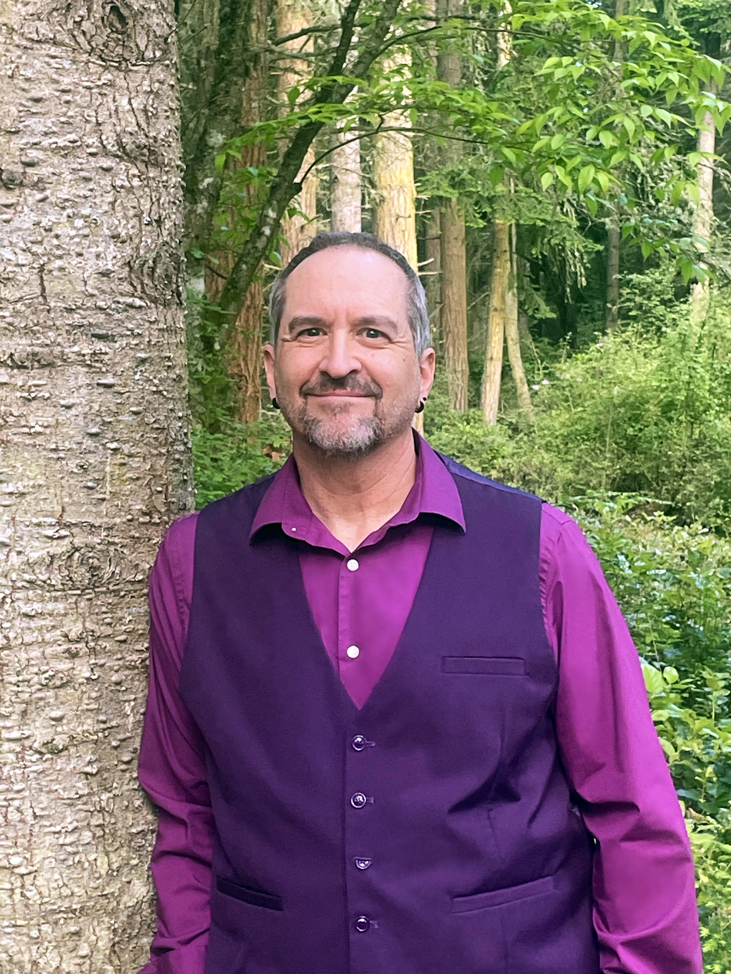 A man with gray hair, a beard, and earrings standing next to a tree in a forested area, wearing a purple shirt and black vest.