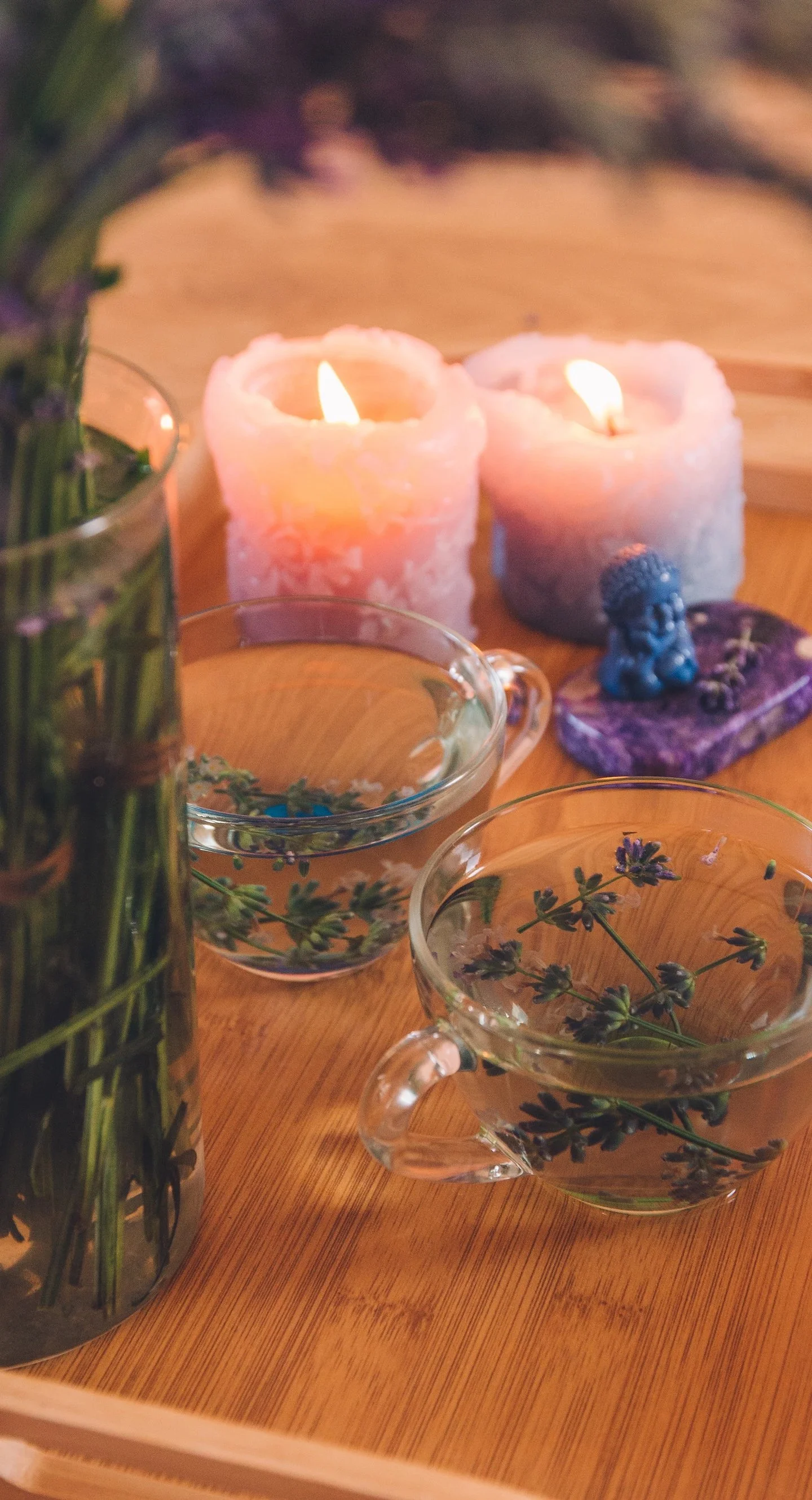 Two lit pink candles, two glass cups with lavender flowers, a purple decorative object on a wooden tray.