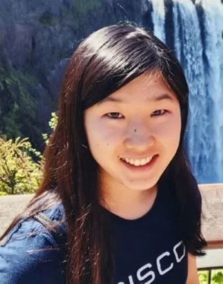 A young woman smiling outdoors near a waterfall, with greenery in the background.