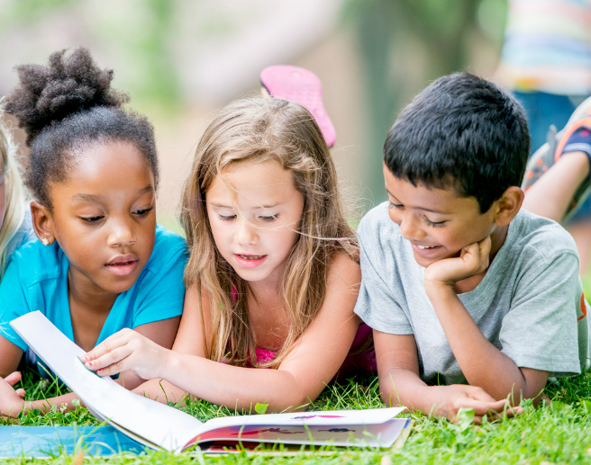 Three children lying on grass outdoors, looking at a book together.